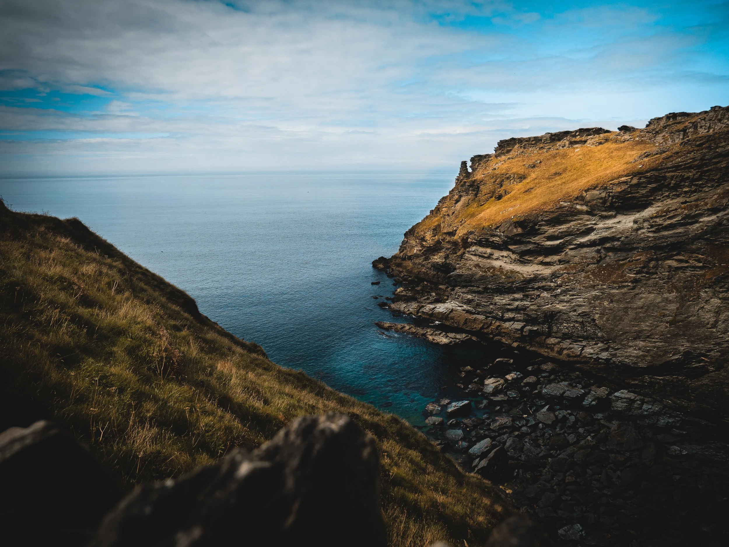 Cliffs overlooking the ocean with a grassy slope on the left and rocky terrain on the right, under a partly cloudy sky.
