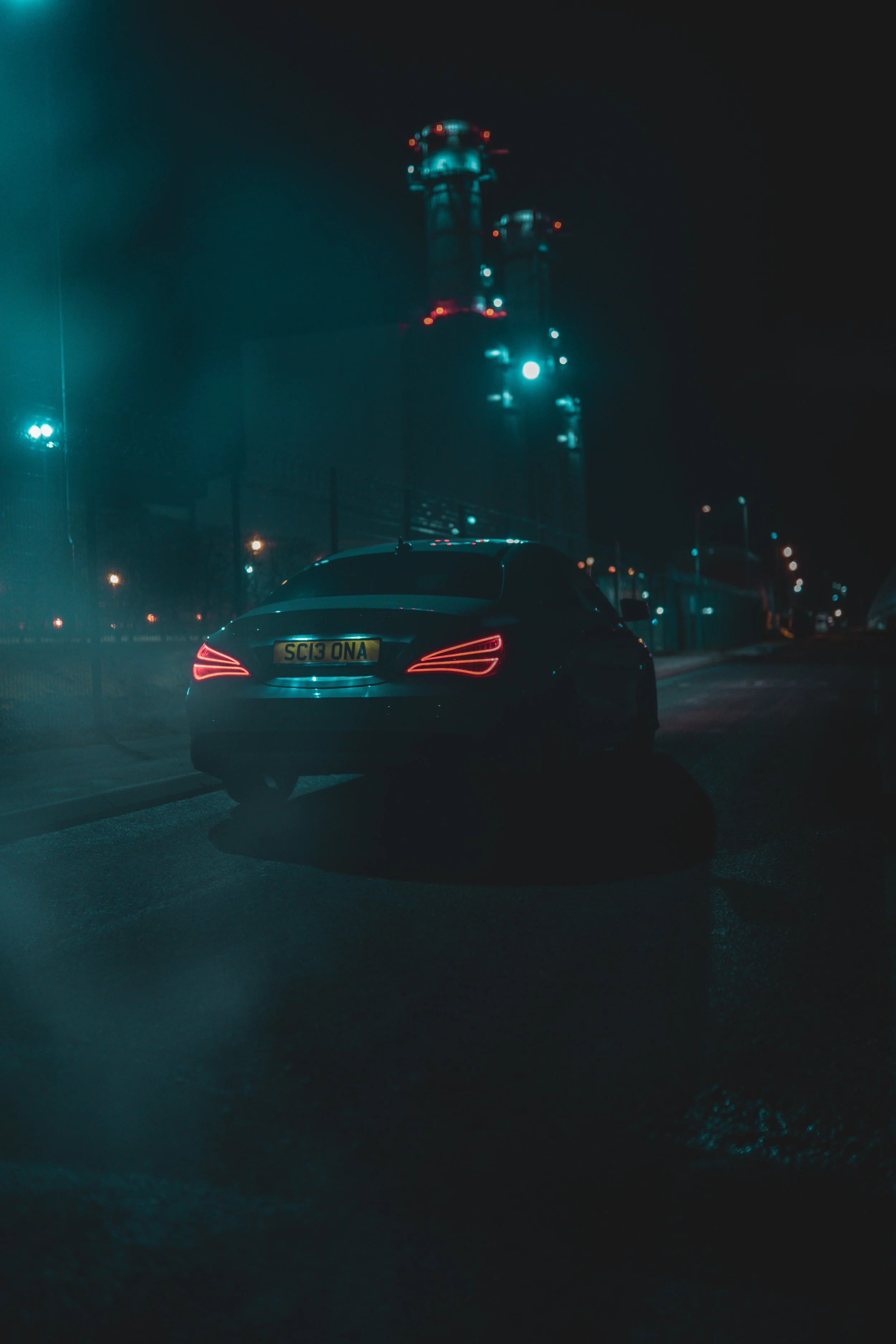 A dark night scene with a black car parked on the street near industrial towers illuminated by teal lights.