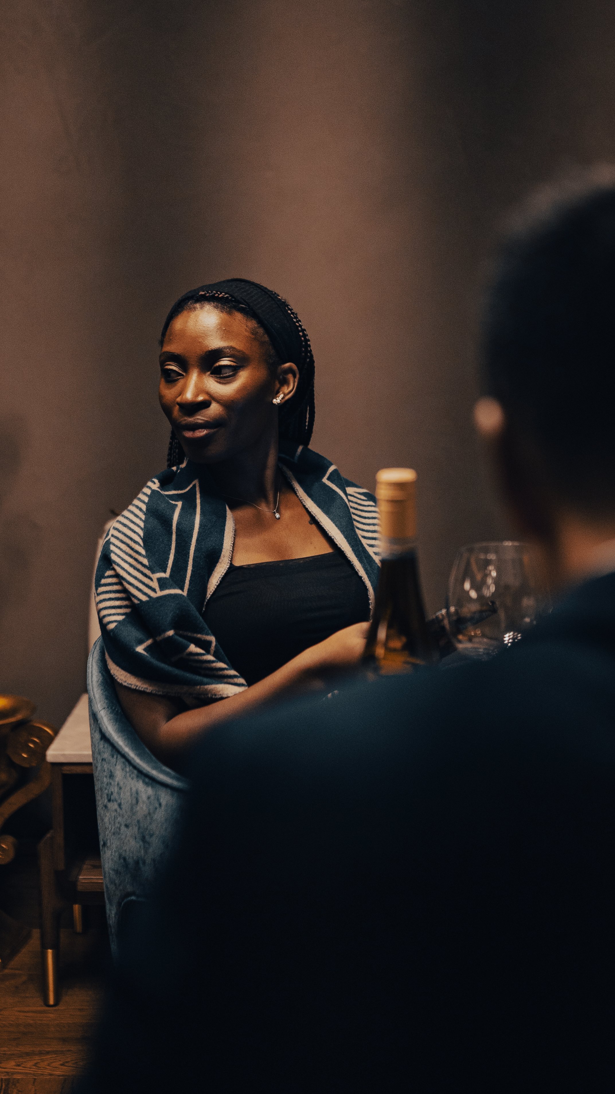 A woman with braided hair wearing a black top and a patterned shawl, sitting at a table with wine glasses and a bottle, in a dimly lit setting.