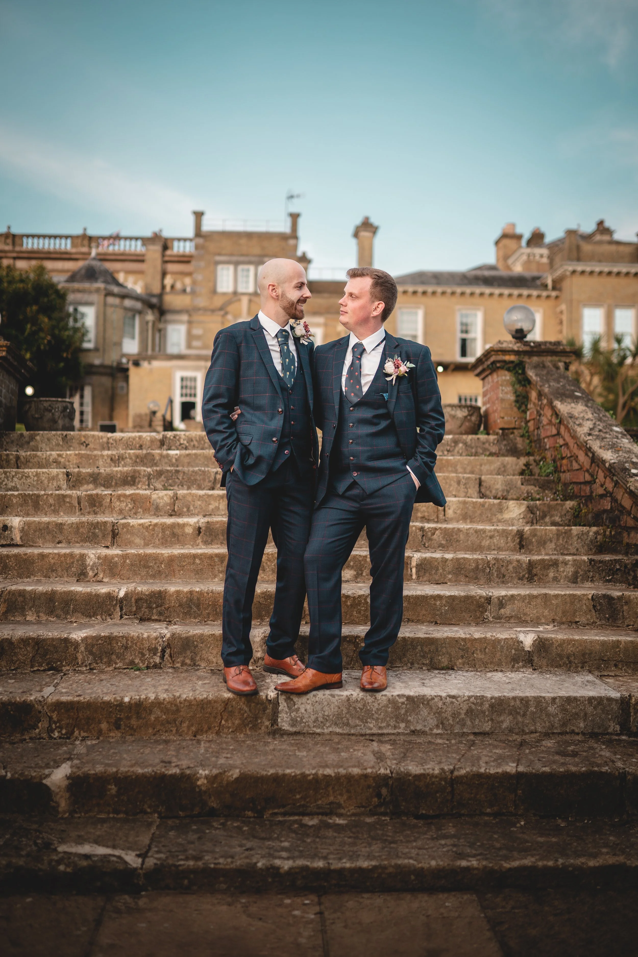 Two men in matching plaid suits standing on stone steps, celebrating a wedding outside. They are smiling at each other, with flowers pinned to their lapels, in front of a historic building under a cloudy sky.