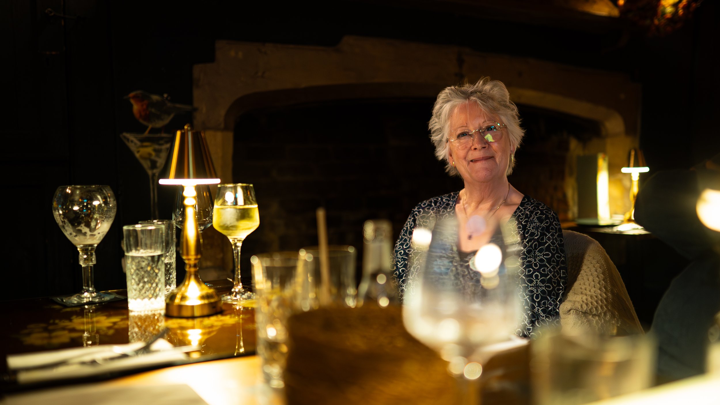 An elderly woman with glasses smiling, sitting at a dimly lit dining table with glasses of wine and water, in a cozy restaurant with a brick fireplace in the background.