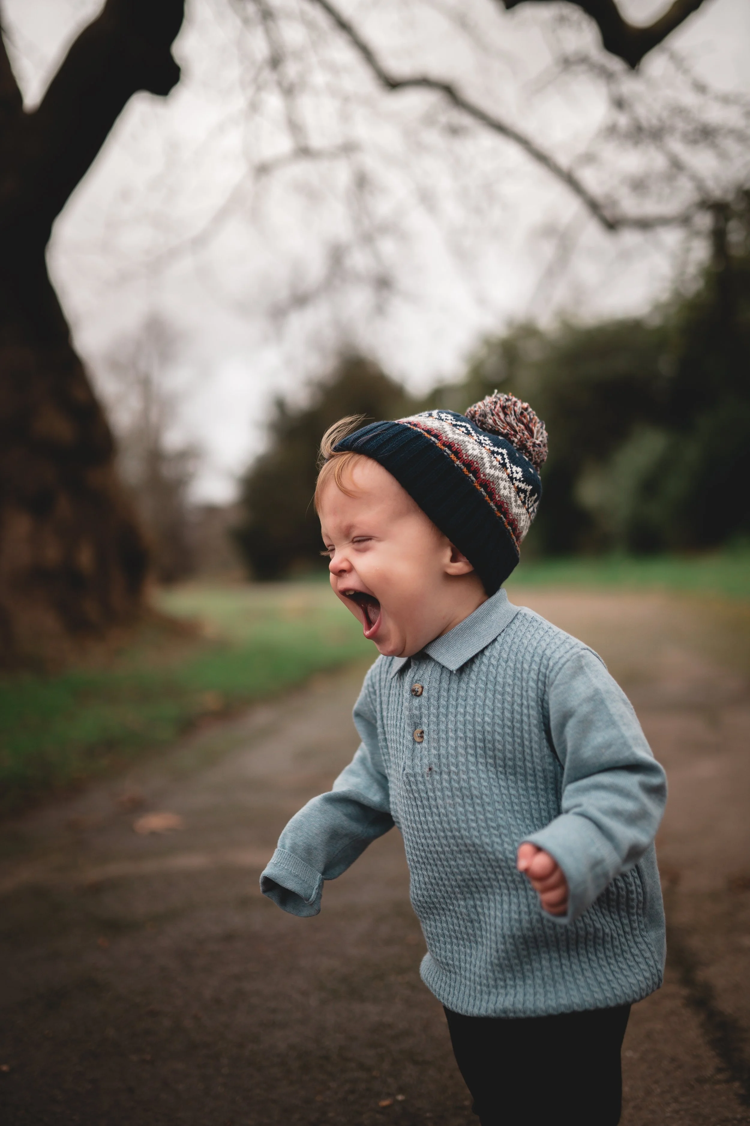 A young boy in a knit hat and gray sweater is outside on a dirt path, laughing or yelling with his mouth open, in a park or wooded area with overcast sky.