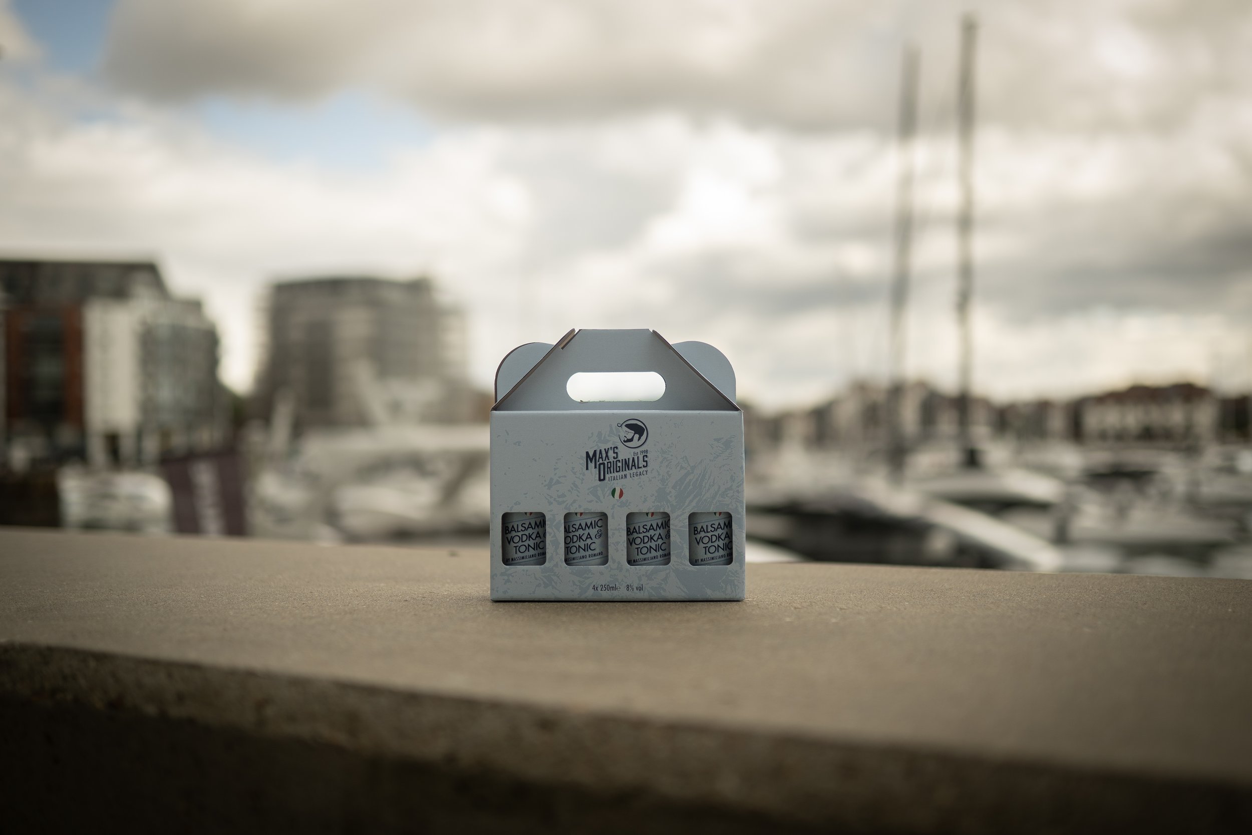 A box of four bottles of Balsamic Vodka Tonic on a concrete surface with a blurred marina and boats in the background.