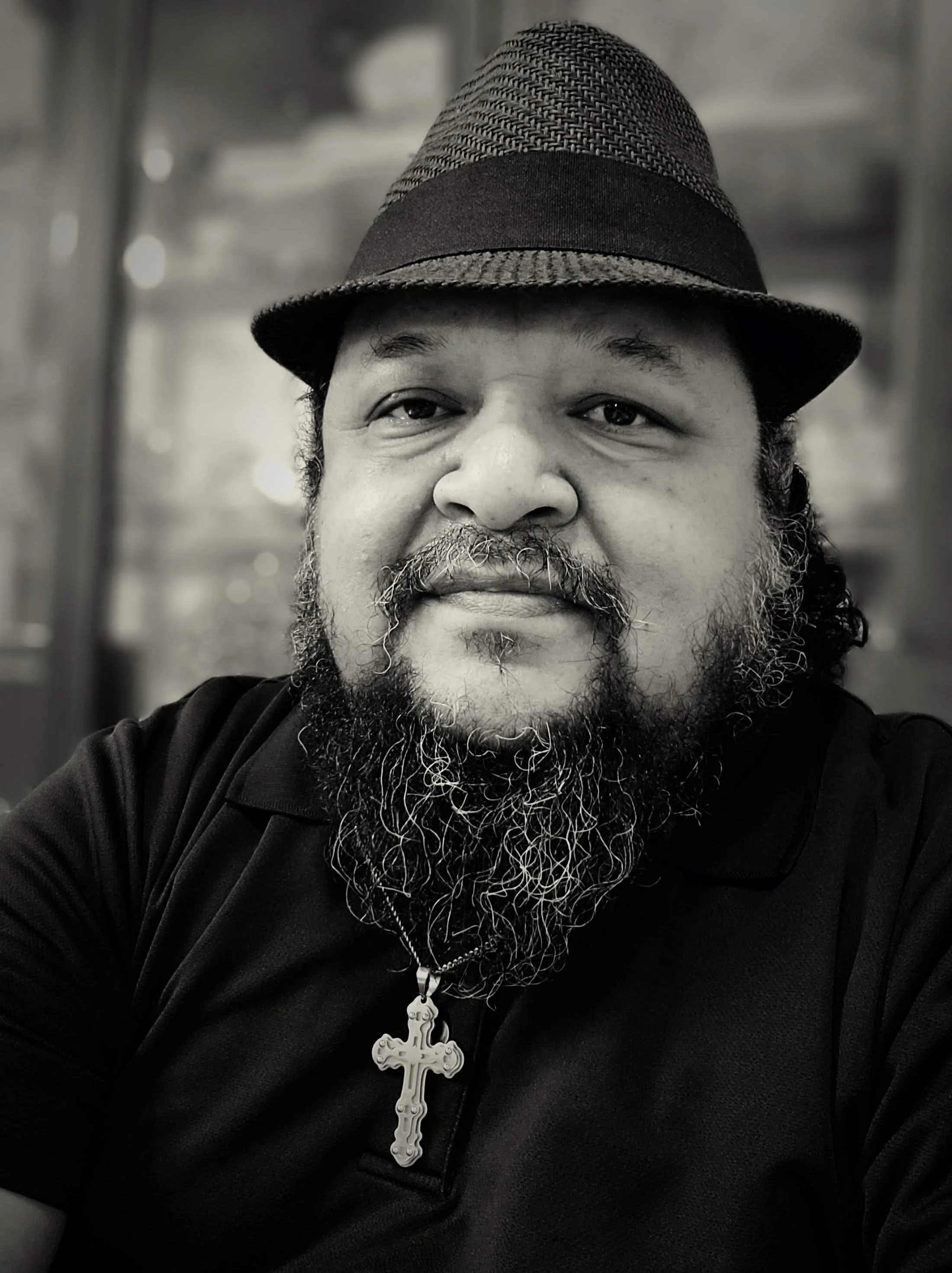 Photo of Rocky, a man with a beard and mustache wearing a fedora hat, a black shirt, and a necklace with a cross pendant.