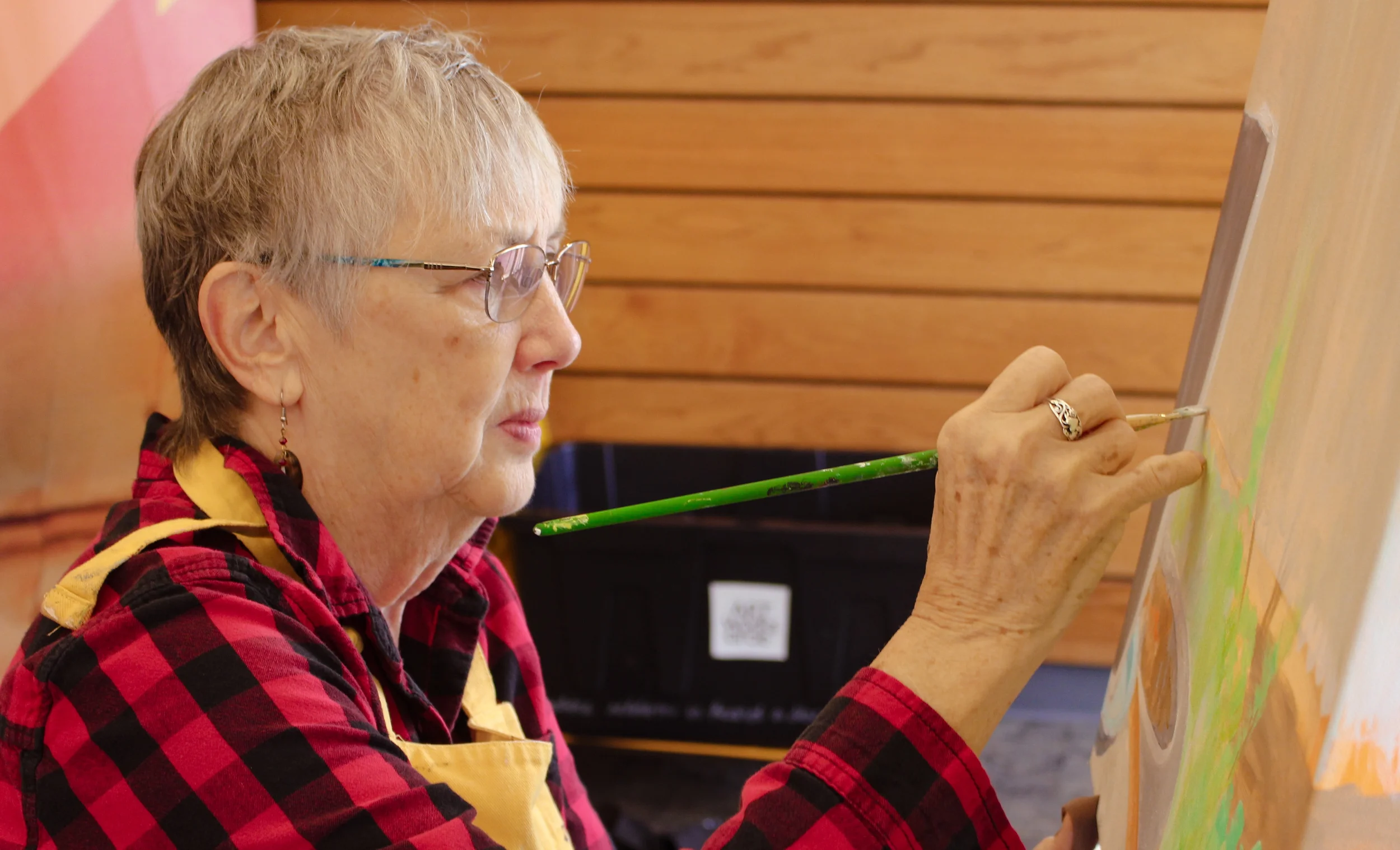 An elderly woman painting on an easel with a paintbrush. She is wearing glasses, a red and black checkered shirt, and a yellow apron. She has short gray hair and earrings.