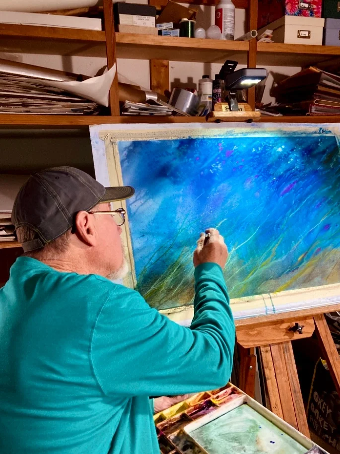 An artist in a teal long sleeve shirt and dark cap painting a colorful abstract watercolor on a large canvas in a cluttered art studio.