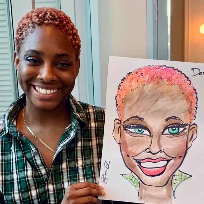 A woman with short, curly reddish hair smiling and holding a colorful caricature drawing of herself, featuring prominent makeup, green eyes, and a wide smile.