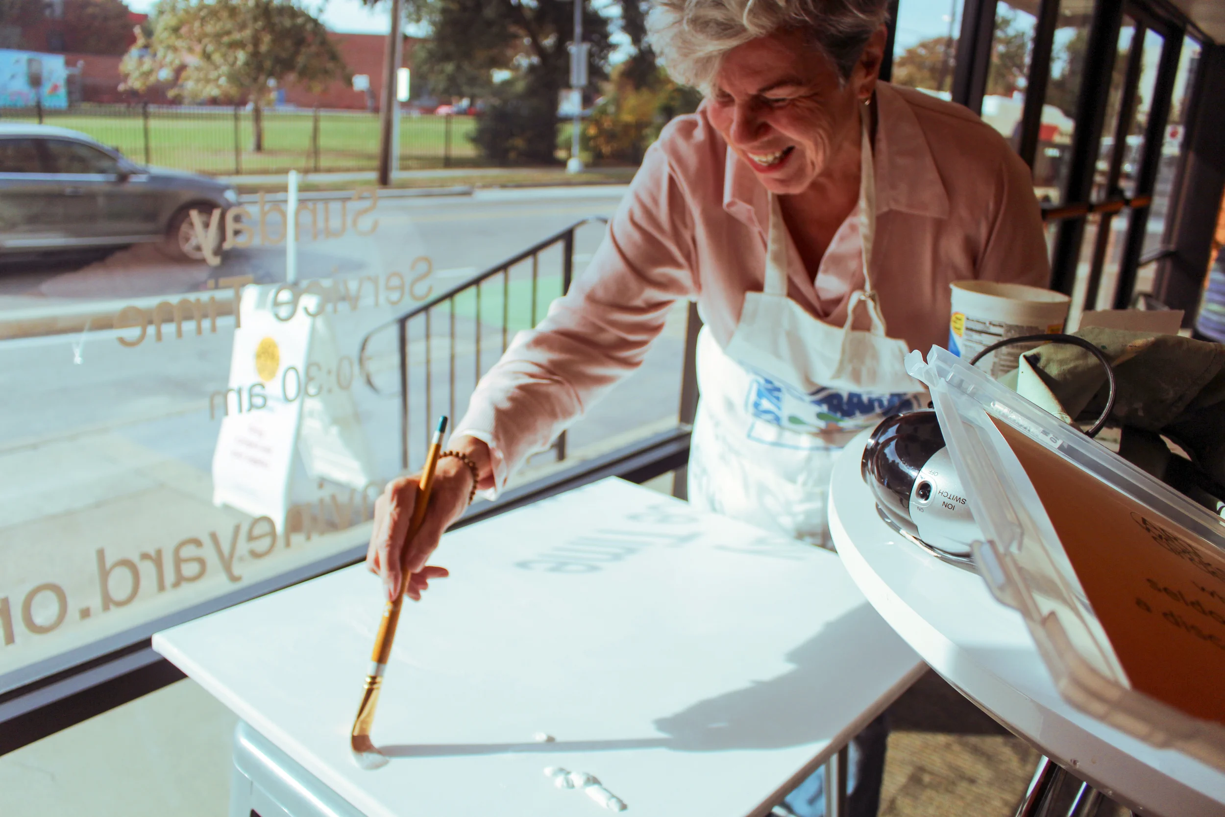 A woman wearing a pink shirt and a white apron is smiling and as she primes a canvas in a sunny room with large windows. Outside is a street with cars and trees.