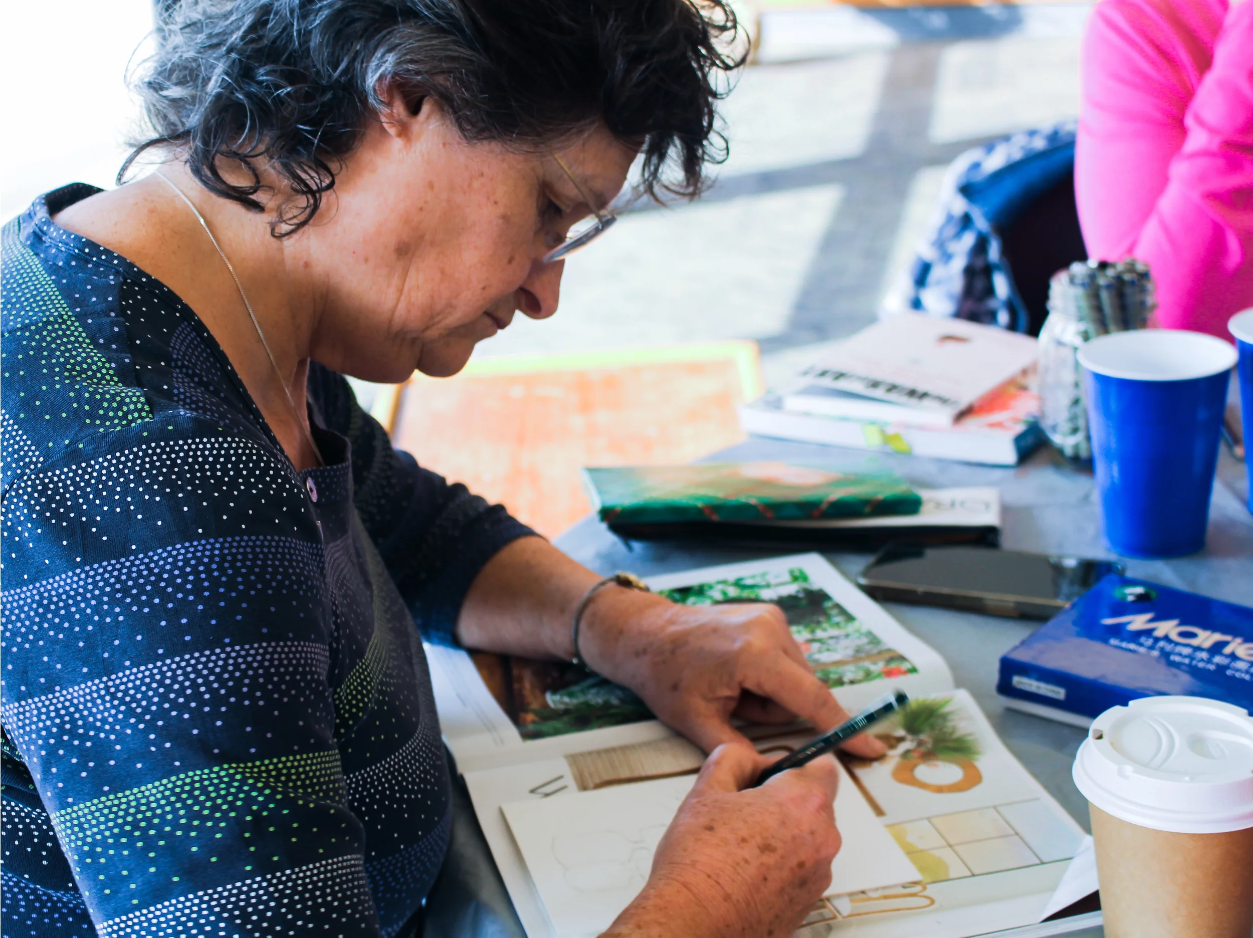 A woman with glasses and curly dark hair is sitting at a table, looking at a book with Christmas-themed images, holding a pen in her hand. The table has various books, a smartphone, a blue box of tissues, a disposable coffee cup, and cups, with anoth