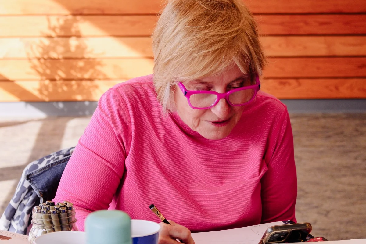 An elderly woman wearing pink glasses and a pink shirt, sitting at a table, looking at her smartphone, with pens and a cup on the table.