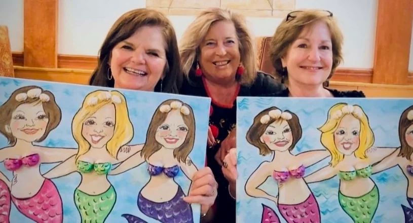 Three women smiling, holding up illustrated caricatures of themselves as mermaids.