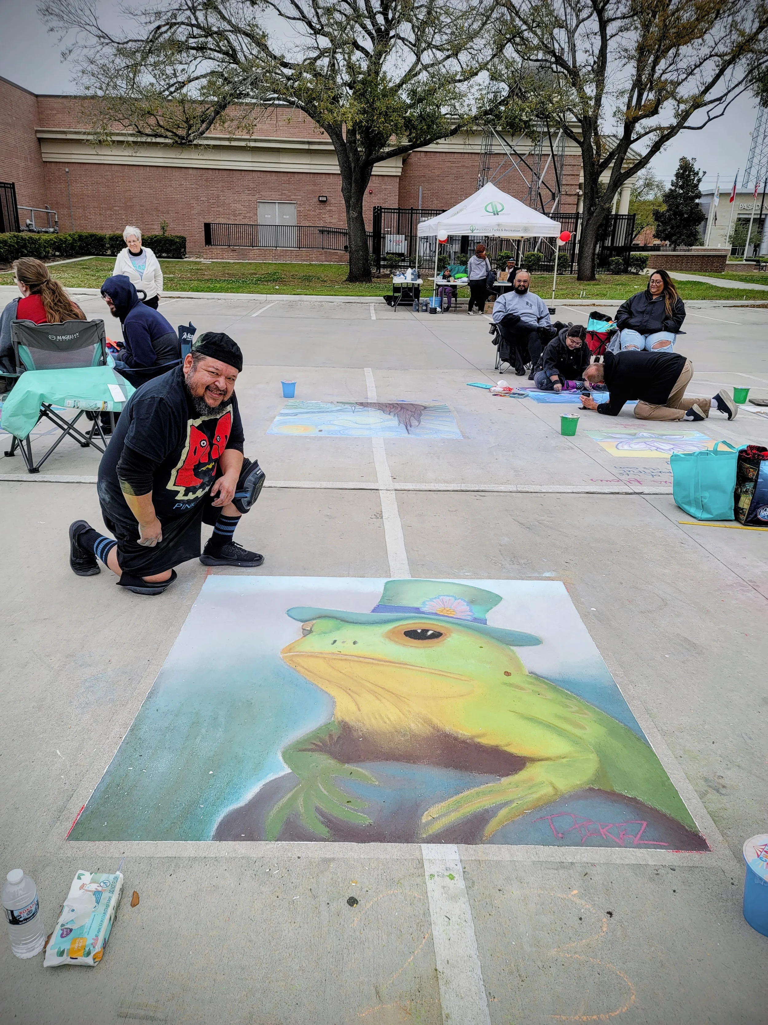 A man kneeling and smiling next to a massive chalk drawing of a frog wearing a hat, in an outdoor parking lot with other artists and onlookers.