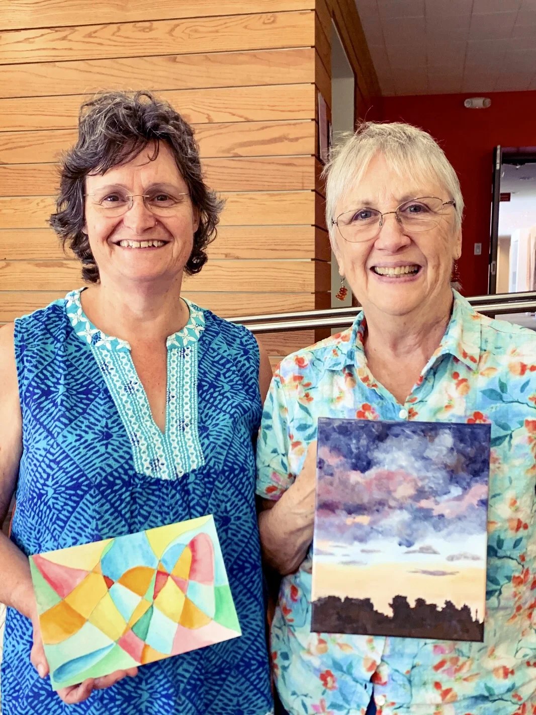 Two smiling women holding colorful paintings, standing in front of a wooden wall, with one painting featuring abstract geometric shapes and the other a landscape with a cloudy sky.