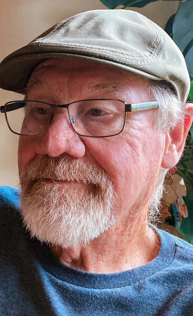 Close-up photo of Mike, an elderly man, wearing a beige cap and glasses with a colorful arm. He has a white beard and mustache, and is dressed in a blue shirt. The background features leafy plants.