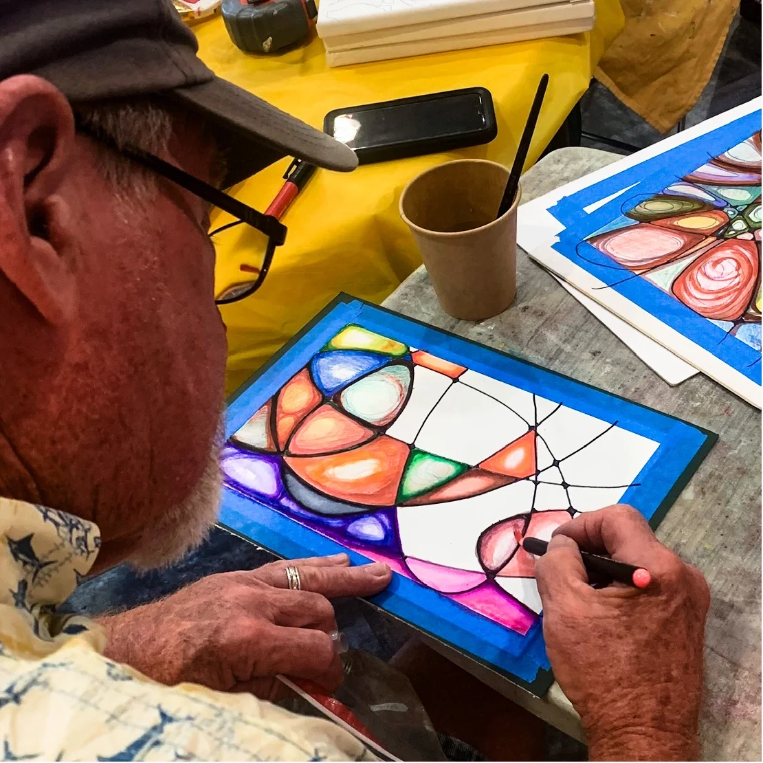 Older man with glasses, wearing a cap and floral shirt, working on a stained glass art piece with colorful glass pieces and black leading, mounted on a board. The table has craft supplies, a cup with straw, and a smartphone.