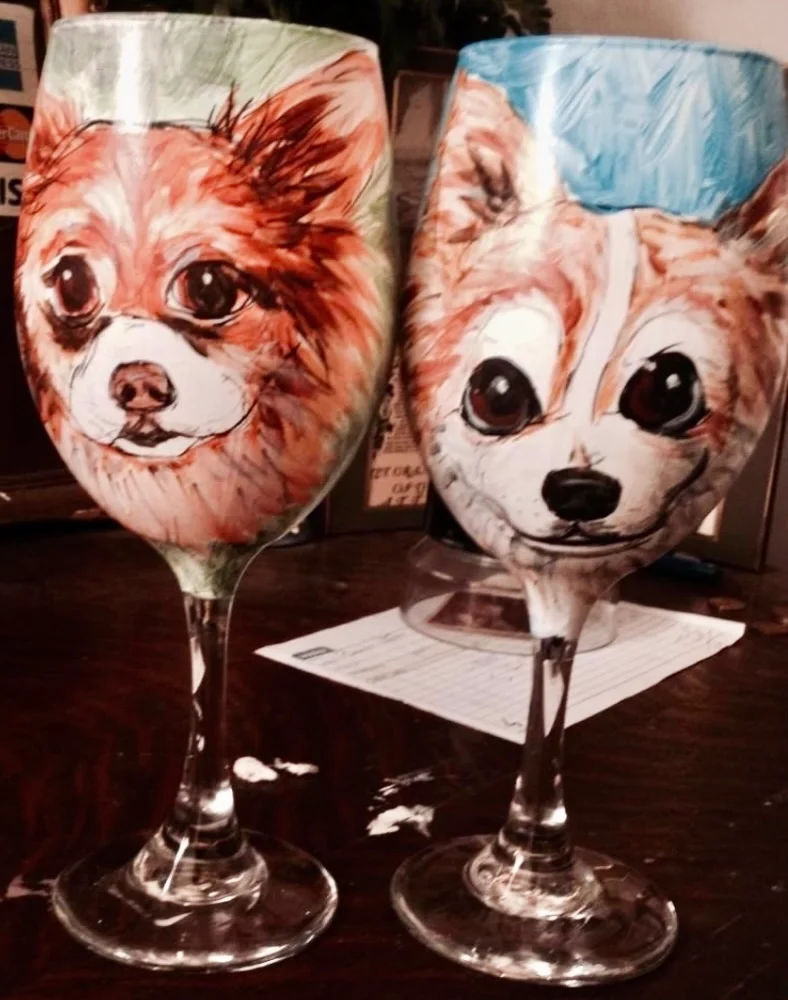 Two wine glasses with hand-painted portraits of cute dogs on them, one with a brown and white dog with big eyes and the other with a similar dog with a blue background.