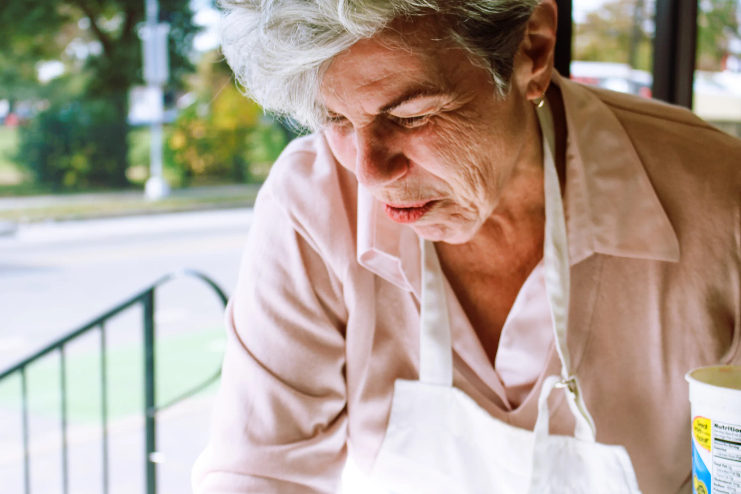 An elderly woman with gray hair wearing a white apron appears to be preparing food indoors with a window showing an outdoor scene of trees and a street in the background.