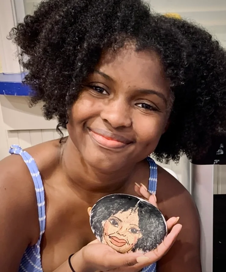 A woman with curly black hair and a friendly smile holding a small painted rock with a portrait of her painted on it.