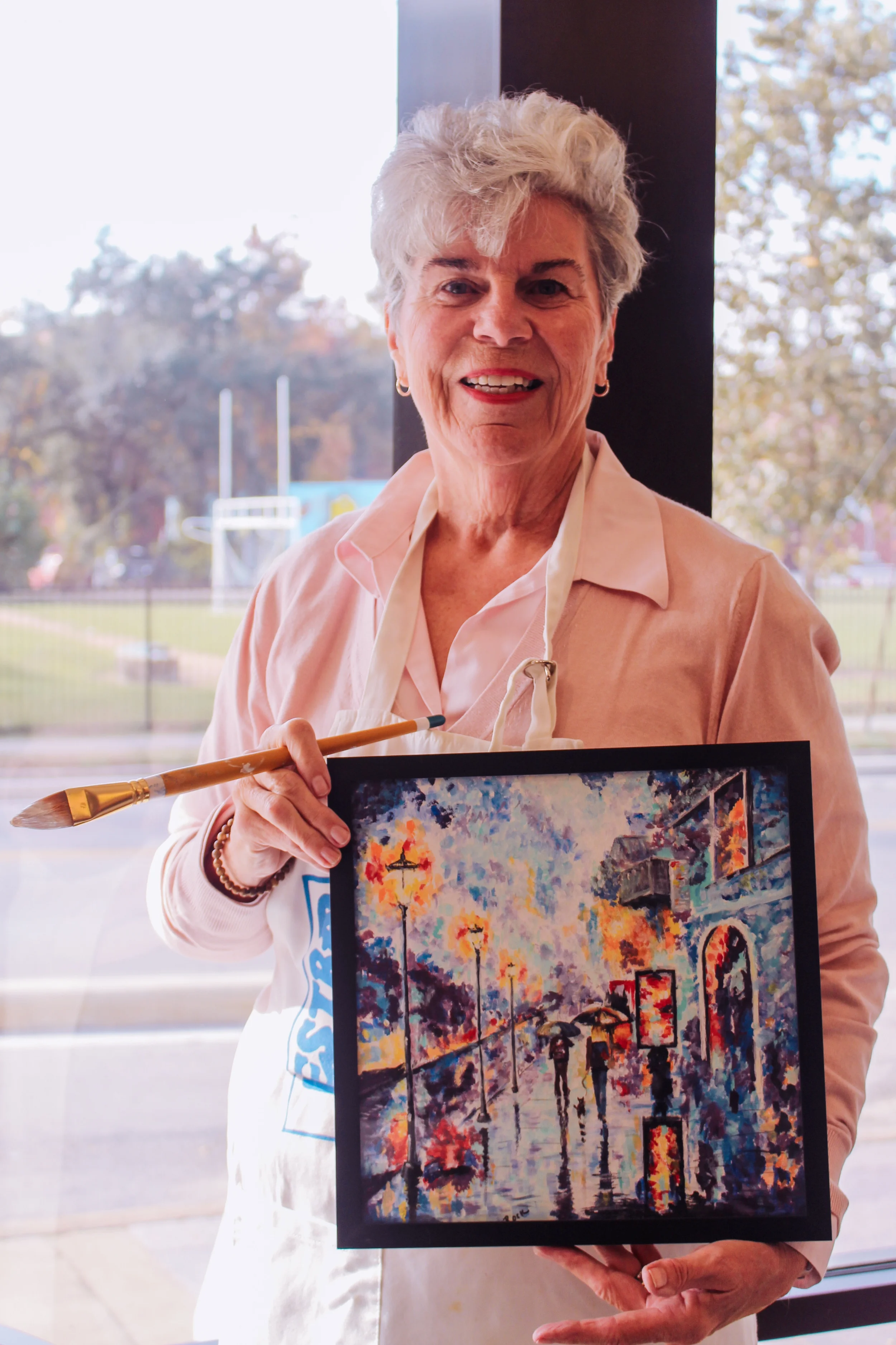 An elderly woman with gray hair smiling, holding a colorful impressionist painting of a city street scene with people walking in the rain, wearing a pink shirt and white apron, standing in front of a window with trees outside.