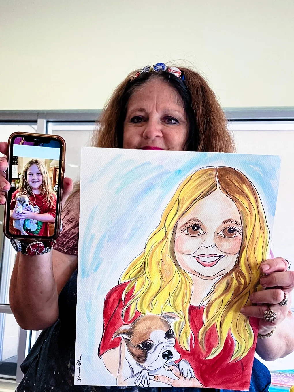 Woman holding a cartoon portrait of a girl with long blonde hair and a small dog, and showing a photo of the girl with the dog on her phone.