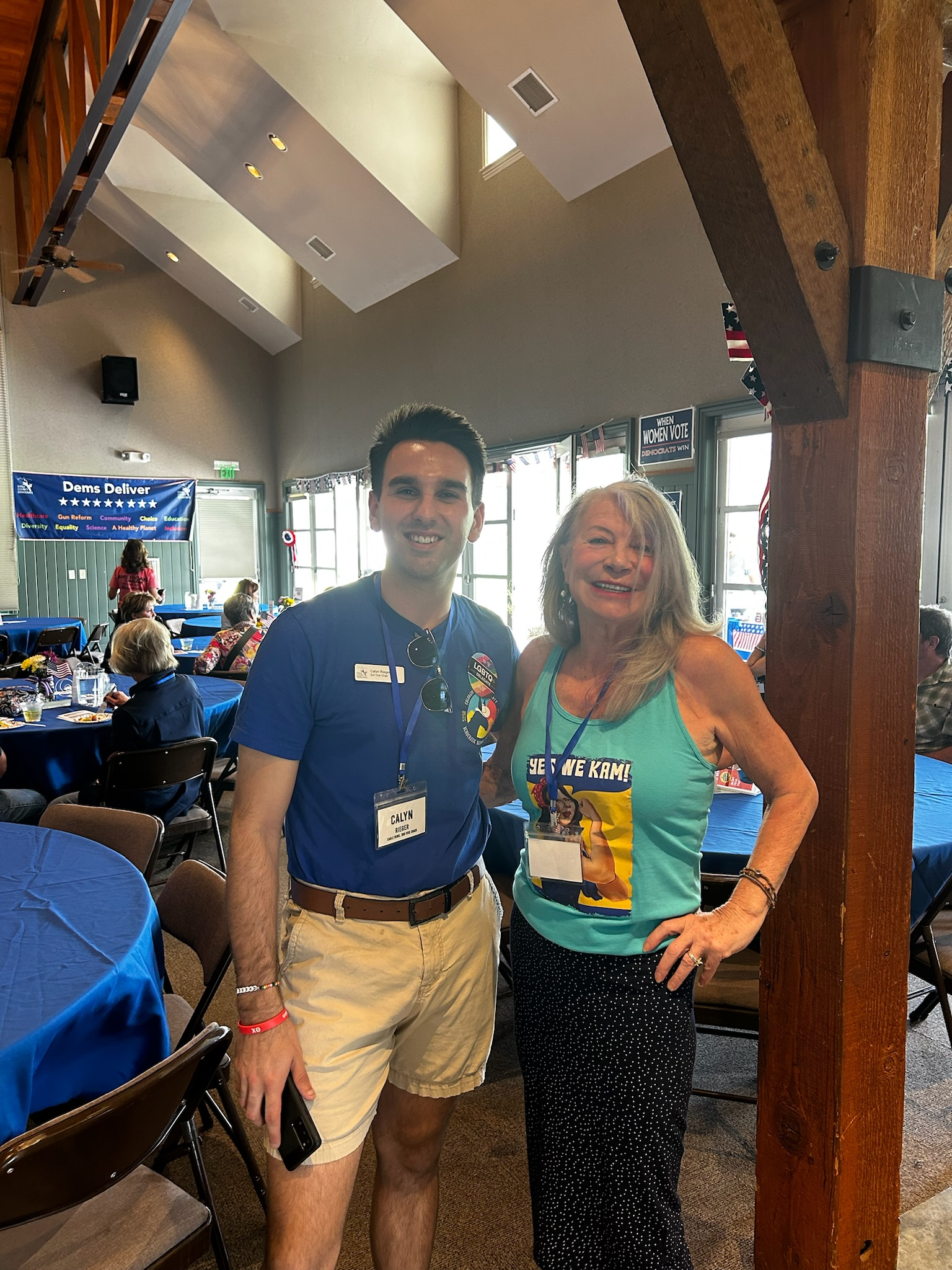 Two people standing inside a decorated event hall, smiling at the camera. The person on the left is a young man wearing a blue shirt, beige shorts, and name tag, holding a phone. The woman on the right is wearing a teal tank top with a superhero comic print, black polka dot pants, and a name badge. The hall has blue tablecloths, guests seated at tables, and political banners and flags hanging around.