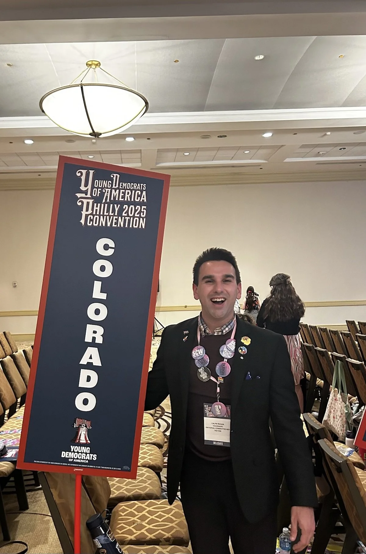 A young man in a black suit and checkered shirt at the Young Democrats of America Philly 2025 Convention, holding a large vertical sign that reads 'COLORADO.' He is smiling, wearing multiple pins, and a delegate badge around his neck. The background shows rows of chairs and a few people.