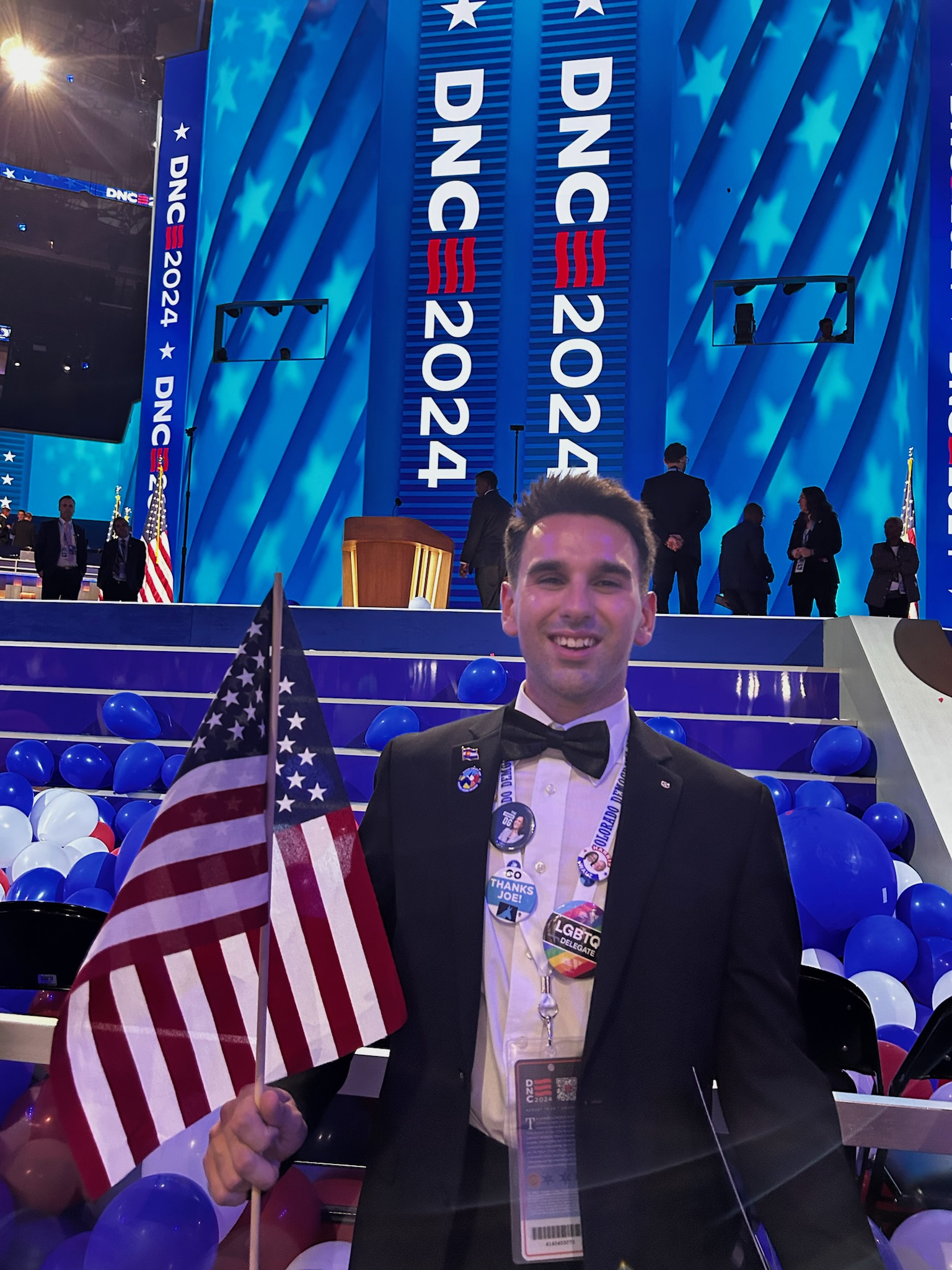 Young man in a tuxedo holding American flag at the 2024 DNC convention with balloons and large screens displaying 'DNC 2024' in the background.