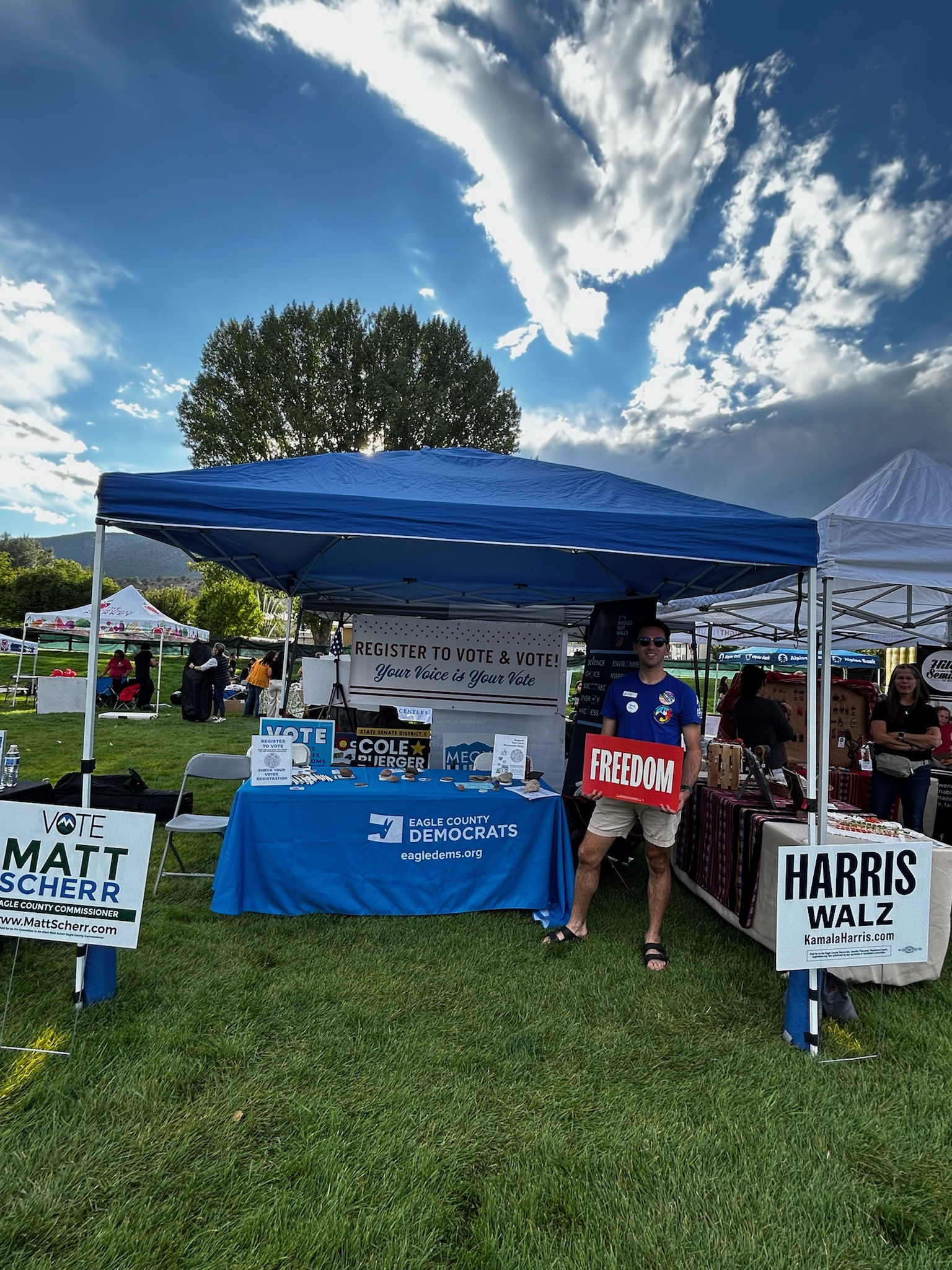 People at an outdoor voting booth and campaign booths, with a man holding a red sign that says "FREEDOM," under a partly cloudy sky and large tree in the background.