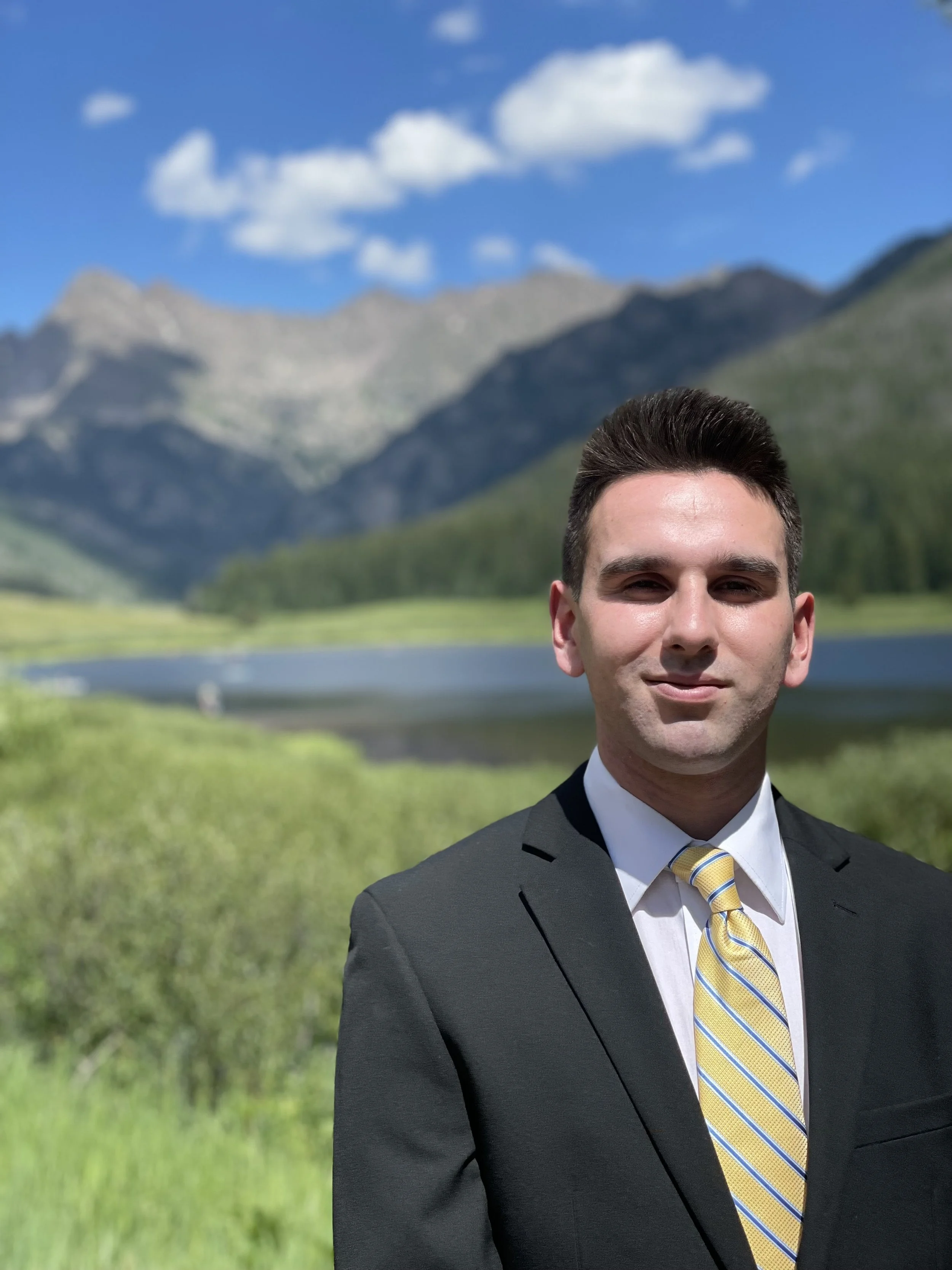 A man in a black suit, white shirt, and yellow striped tie standing outdoors in front of a scenic mountain landscape with a lake, green grass, and a partly cloudy blue sky.