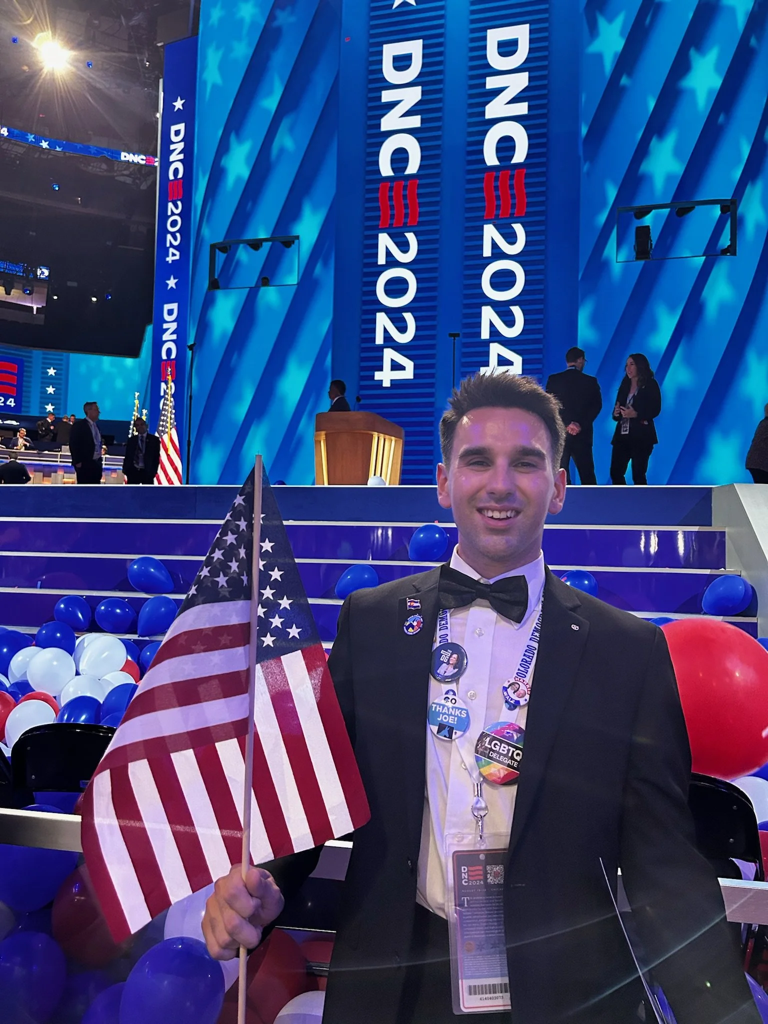 A smiling young man in a tuxedo holding an American flag at the DNC 2024 event, with balloons and a large digital stage displaying 'DNC 2024' in the background.
