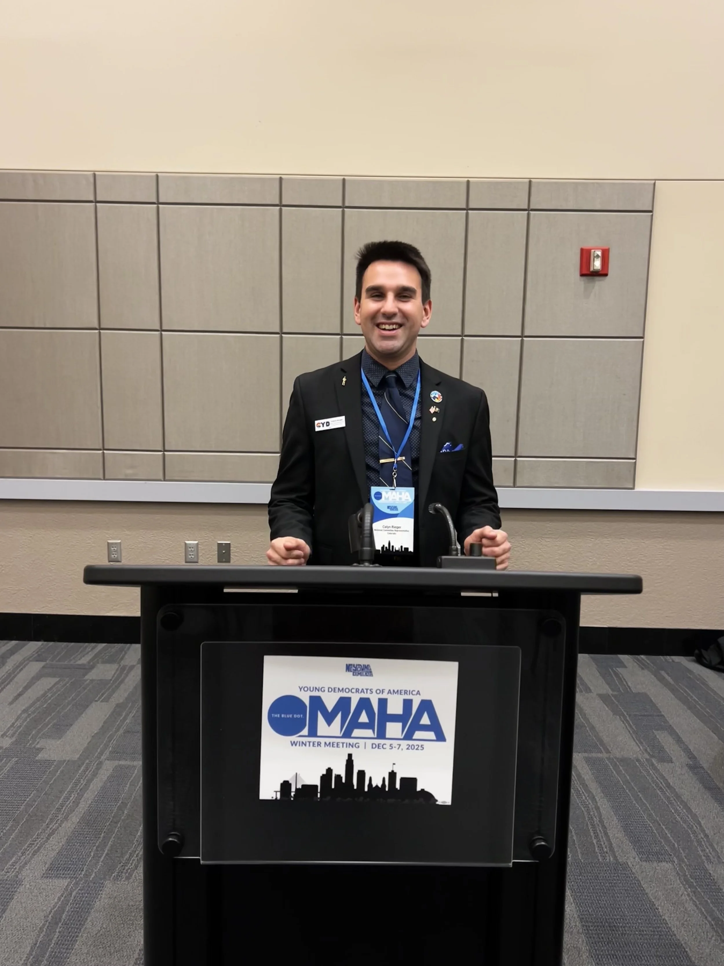 Young man standing at a podium with a 'Young Democrats of America' display, smiling at an indoor conference.