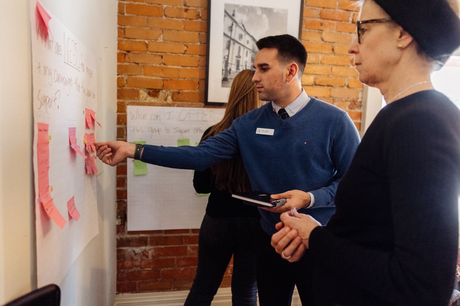 Two women and a man are standing near a whiteboard with pink sticky notes, engaged in a discussion or presentation. The man is pointing at the sticky notes and holding a notebook, while one of the women looks at the board. The room has a brick wall and framed pictures.