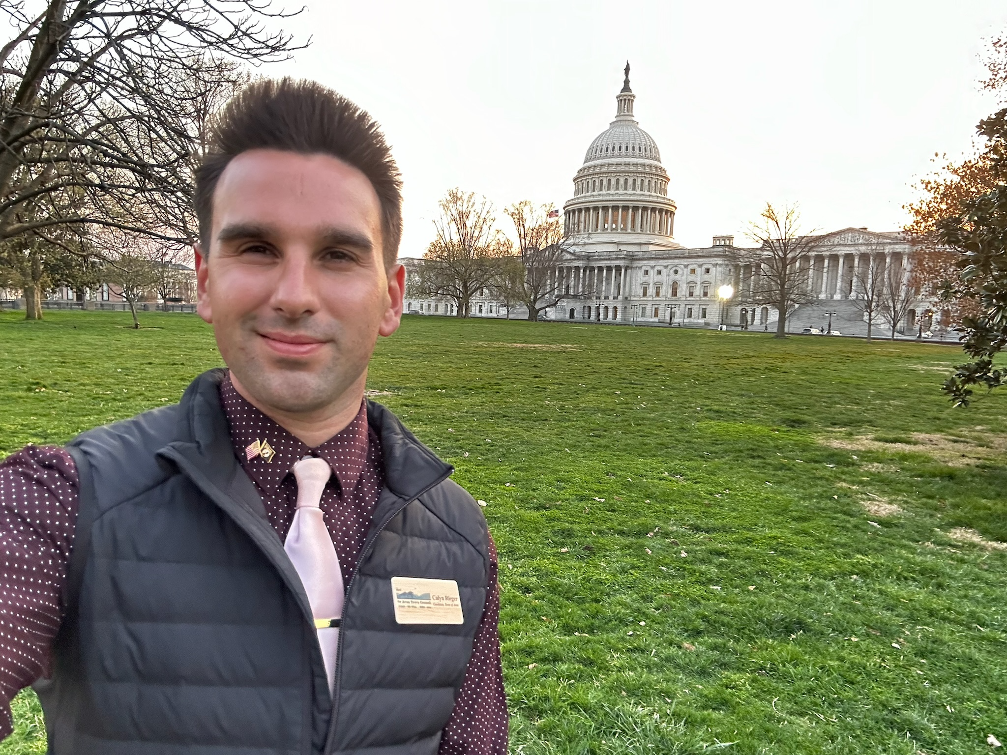A man taking a selfie in front of the U.S. Capitol building on a grassy area with trees and a pathway. The man is wearing a black vest, a maroon polka-dot shirt, a light pink tie, and has a name tag on his vest.