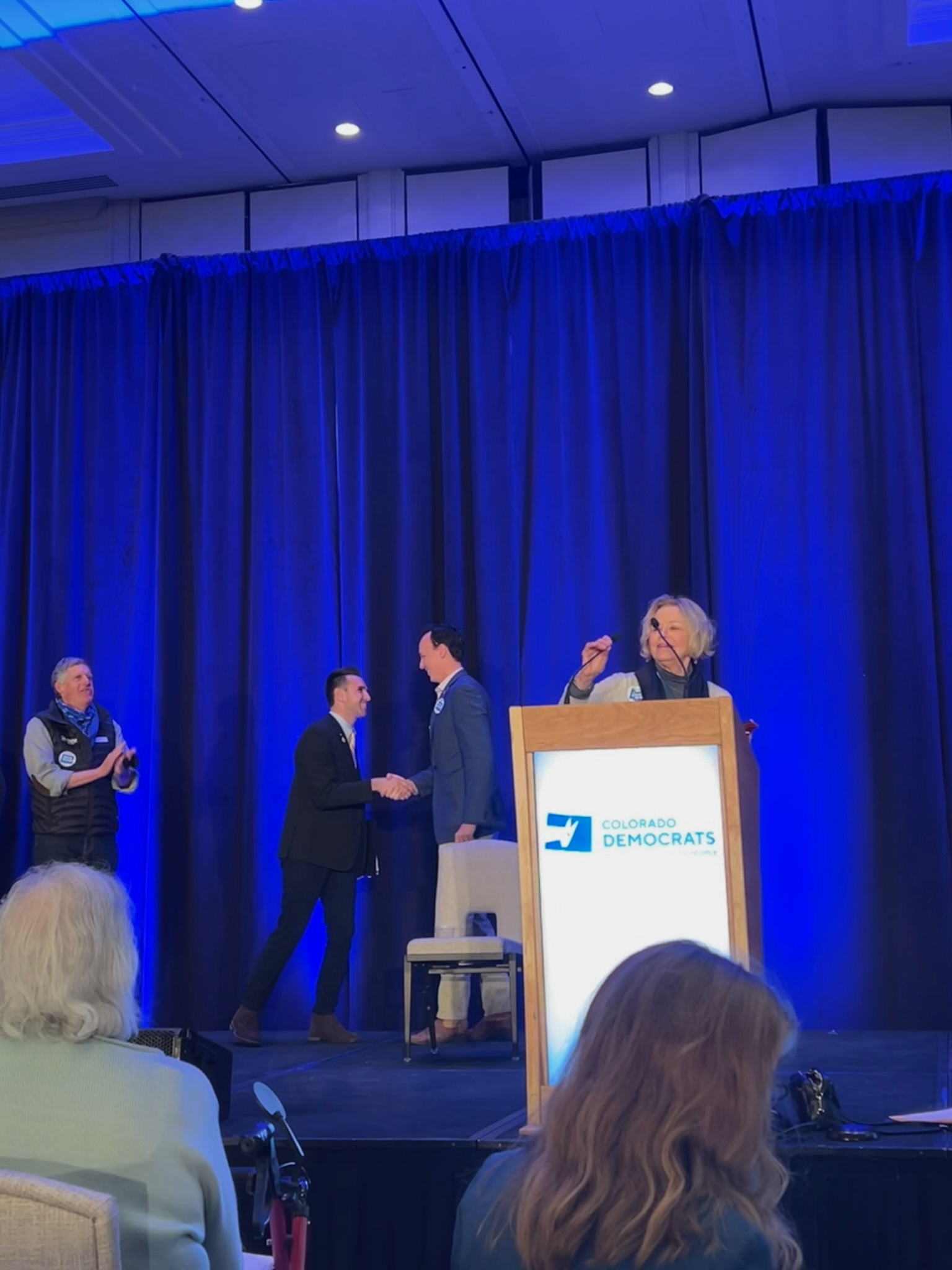 A stage with a blue curtain backdrop at a Colorado Democrats event. Two men in suits are shaking hands at center stage, with a woman standing behind a podium displaying the Colorado Democrats logo. Audience members are visible in the foreground.