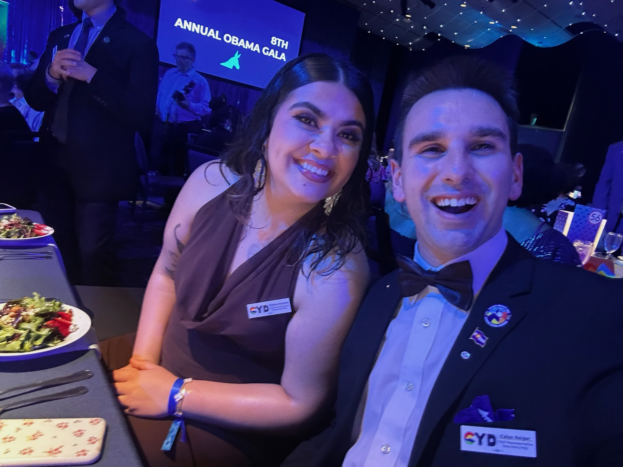 Two people dressed in formal attire taking a selfie at the 8th Annual Obama Gala with a large screen behind them displaying event details. The woman has dark, wavy hair and the man has short hair, both smiling.