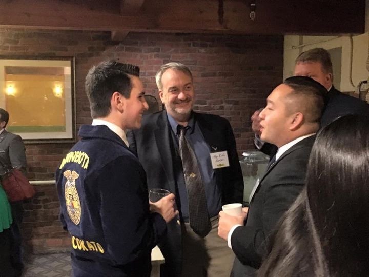 Four people, are engaged in conversation at a social event in an indoor space with brick walls. One man is wearing a suit and tie, another man is dressed in a blazer, and one woman is partially visible with long dark hair. The atmosphere is friendly and professional.