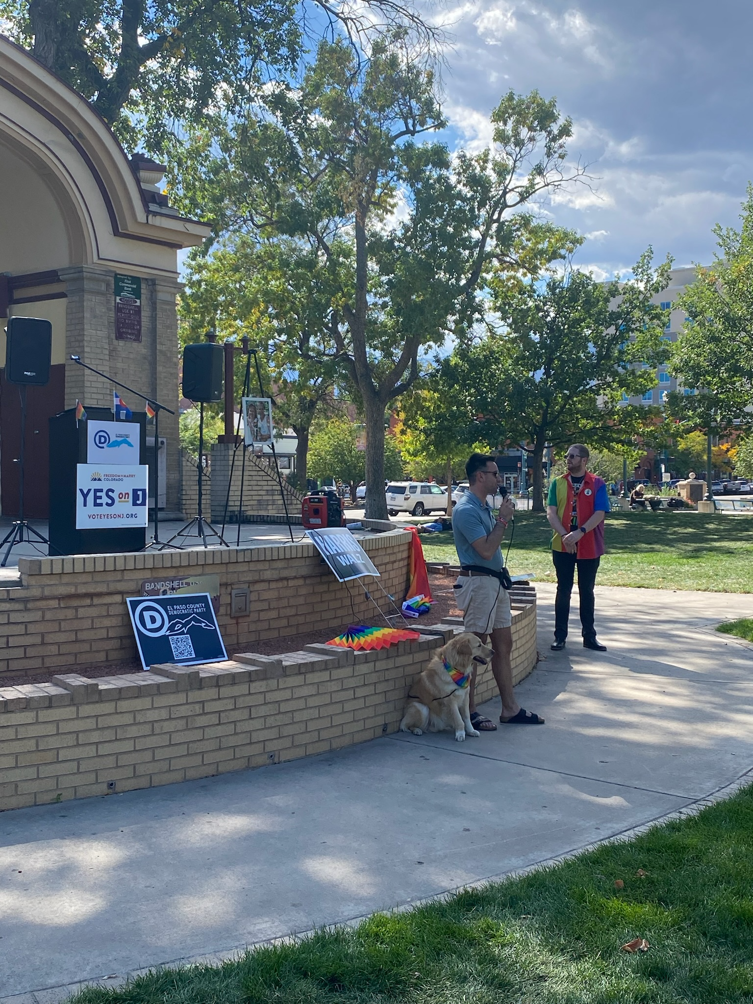 People attending an outdoor event on a park stage, with signs and banners supporting Democratic political campaigns, including a poster that reads 'YES on J' and a QR code, a dog with a rainbow-colored bandana, and trees in the background.