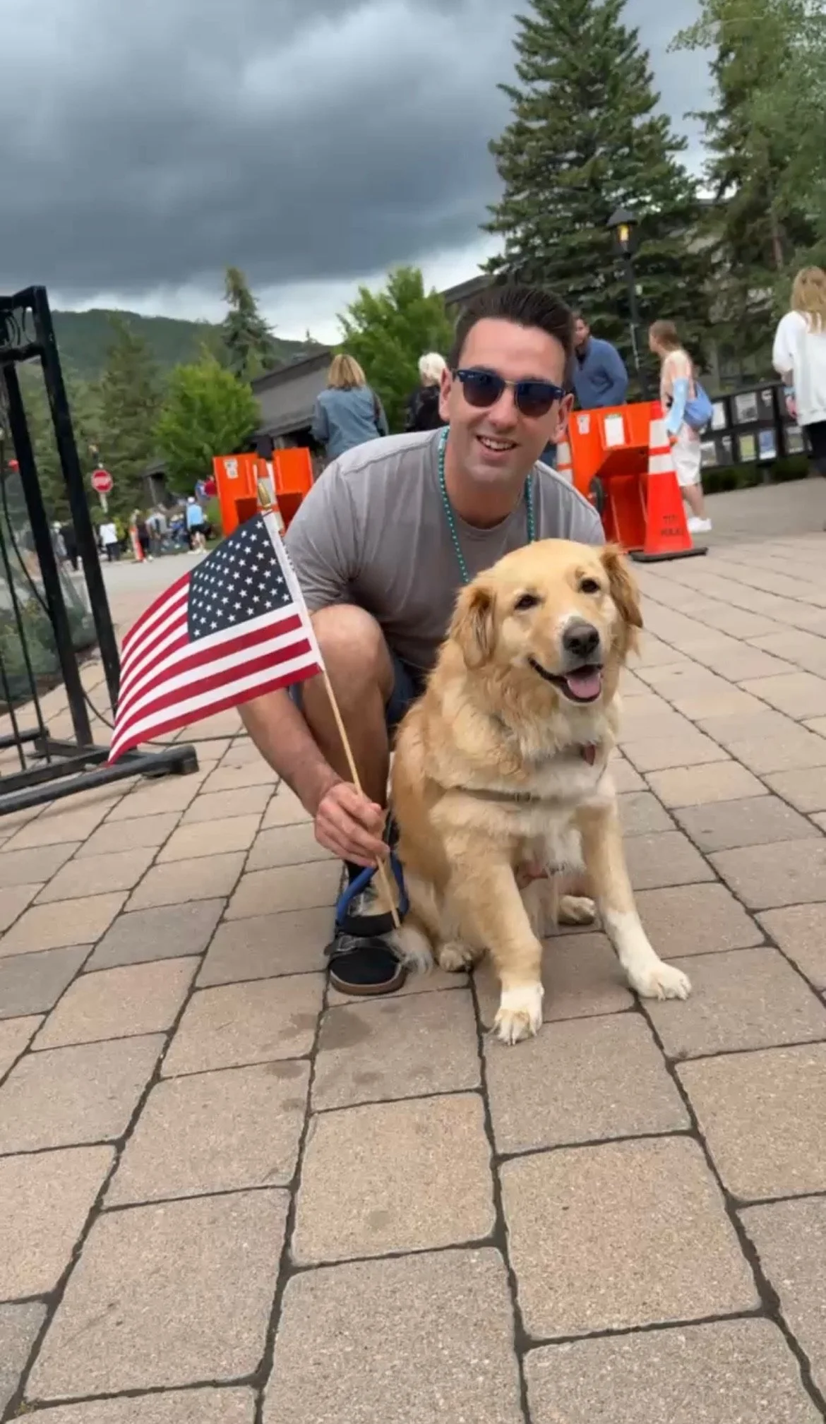 A man kneeling next to a happy golden retriever mix dog on a paved street, holding an American flag, with trees and cloudy skies in the background.