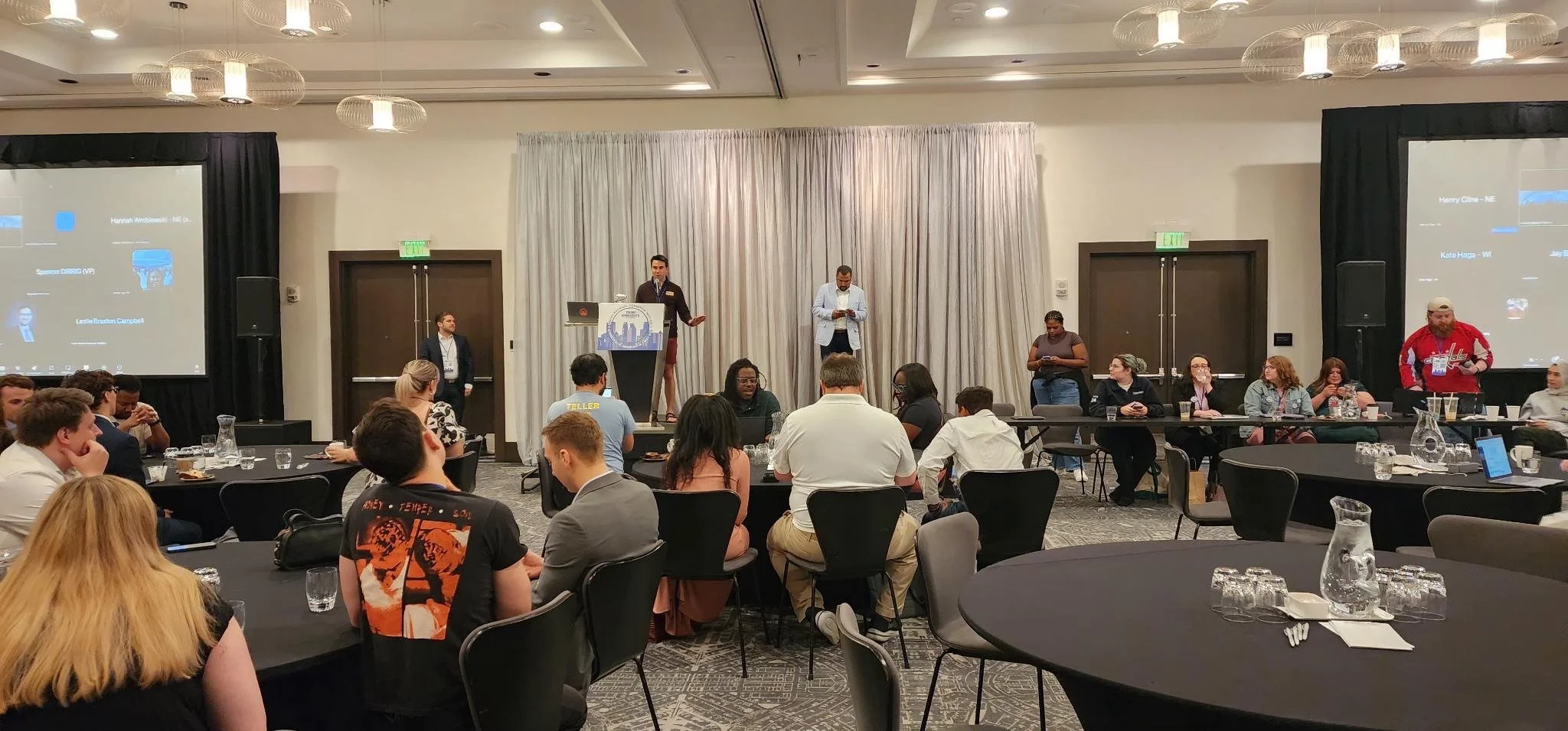 Conference room filled with round tables and attendees listening to a presentation, with two large screens on either side of the stage showing a slide, and a speaker at the podium.