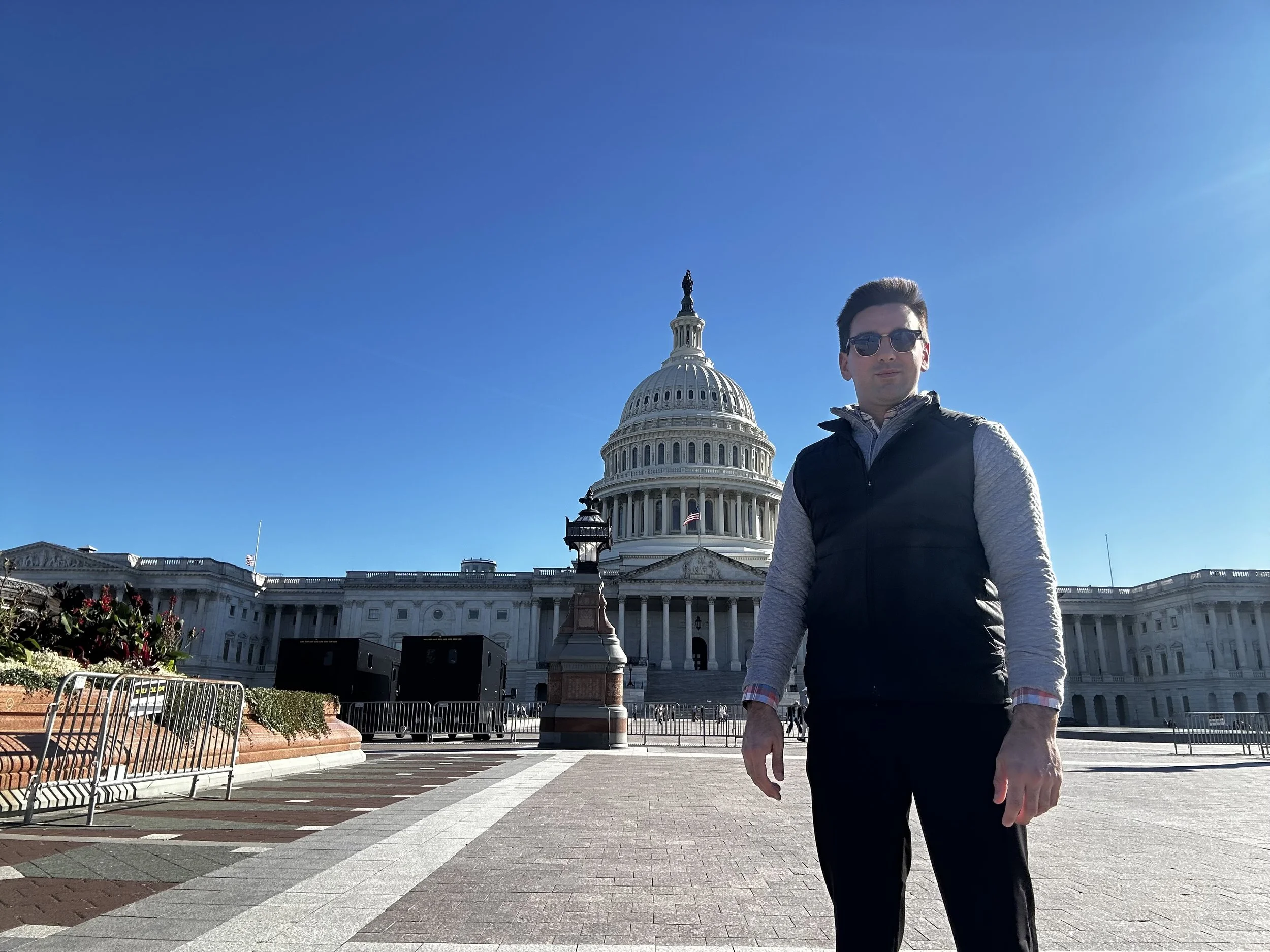 A man wearing sunglasses, a black vest, and gray sleeves stands in front of the U.S. Capitol building on a clear, sunny day.