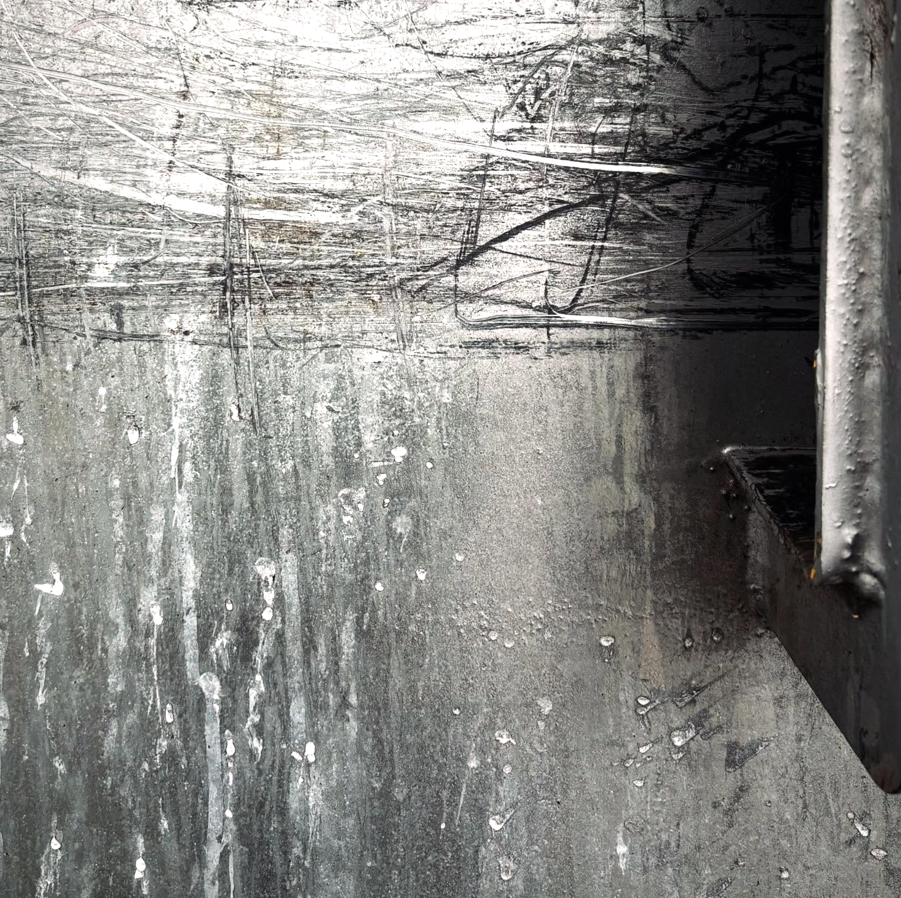 A metallic surface with scratches, scuffs, and water droplets. The surface appears weathered and worn. A part of a metal frame or edge is visible on the right side of the image.