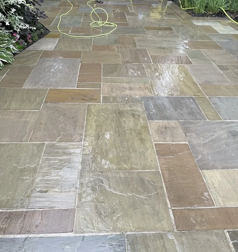 A wet stone patio with various rectangular and square tiles in shades of green, gray, and brown, surrounded by garden plants.