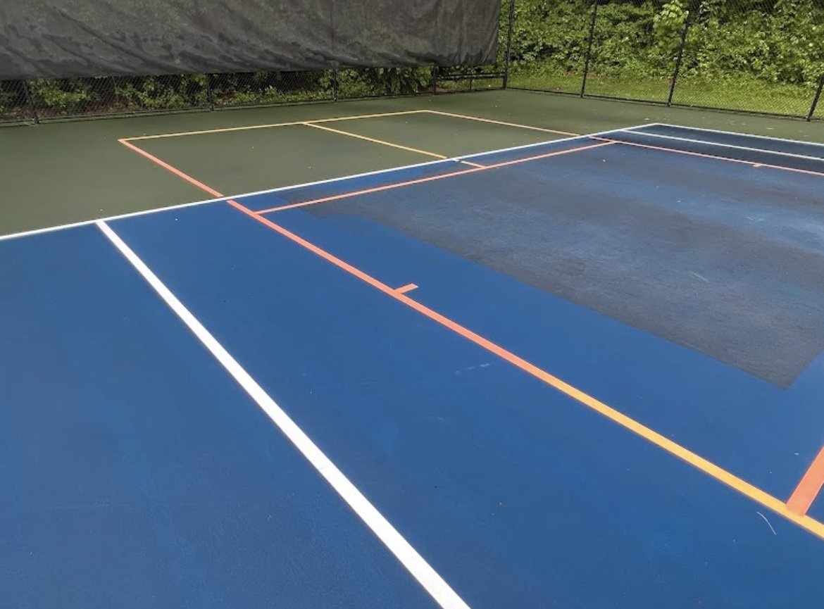 Colorful tennis court with blue, green, and black painted surfaces, surrounded by chain-link fencing and lush green trees.