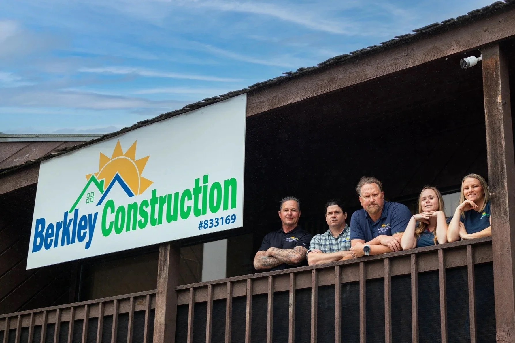 Construction crew standing on a balcony beneath a Berkley Construction sign with a house logo, outdoors against a blue sky with clouds.