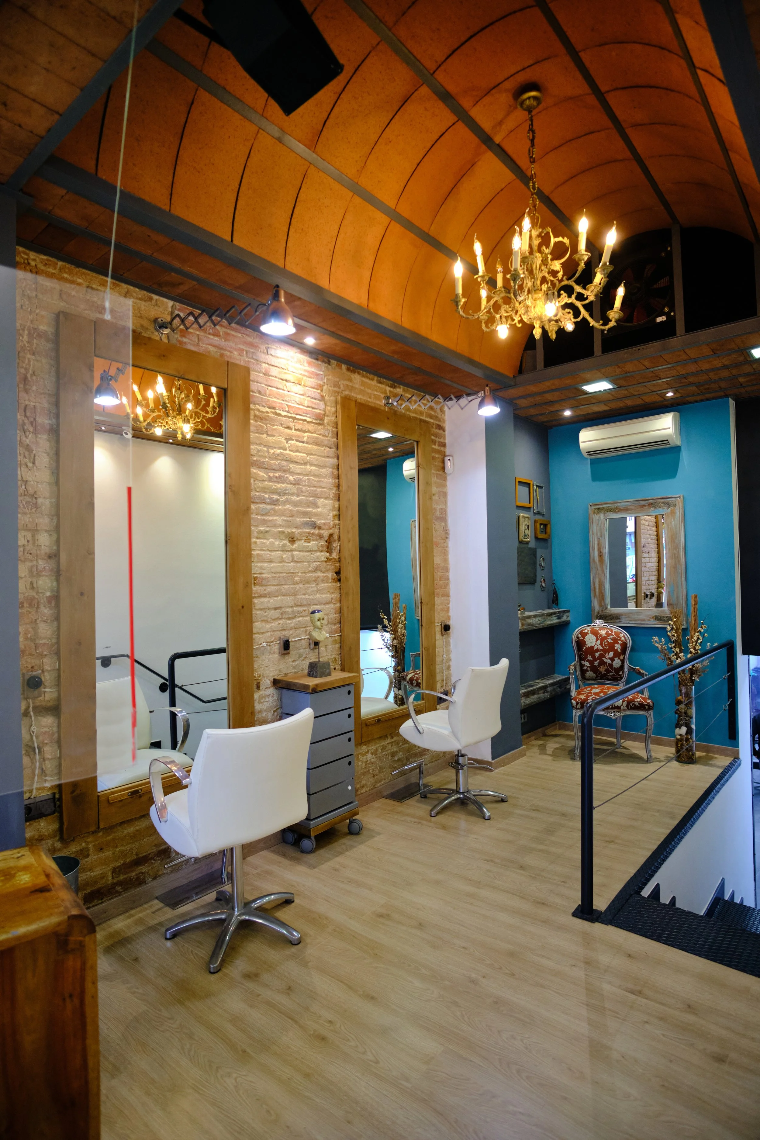 Interior of a modern salon or beauty parlor with wooden floors, exposed brick wall, white salon chairs, large mirrors framed in wood, and a chandelier hanging from the wooden vaulted ceiling. There are also smaller ceiling lights and an air conditioning unit on the blue wall.