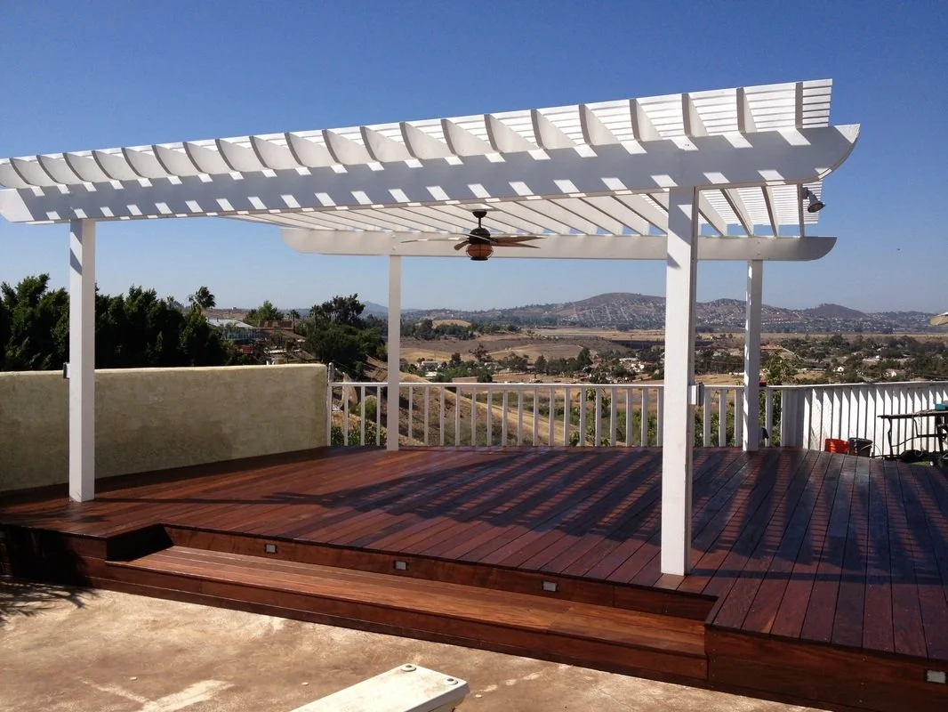 Outdoor patio with a white pergola, wooden deck, ceiling fan, white railing, and scenic view of hills and fields in the background.