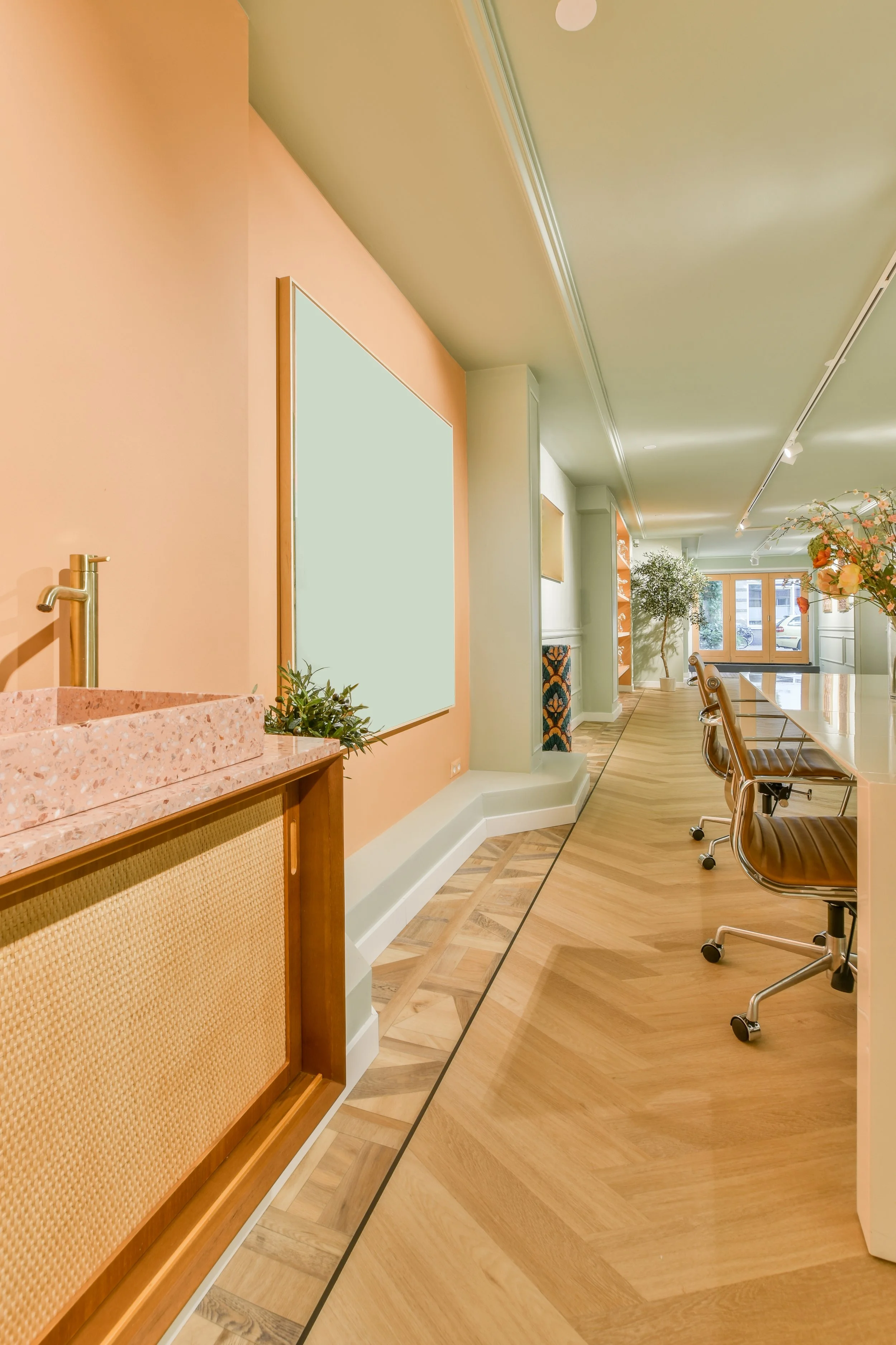 Interior of a modern office or cafe with wooden flooring, a pink wall with a cork panel, green walls, and large windows at the far end. There are brown chairs, a pink terrazzo sink, plants, and floral arrangements.