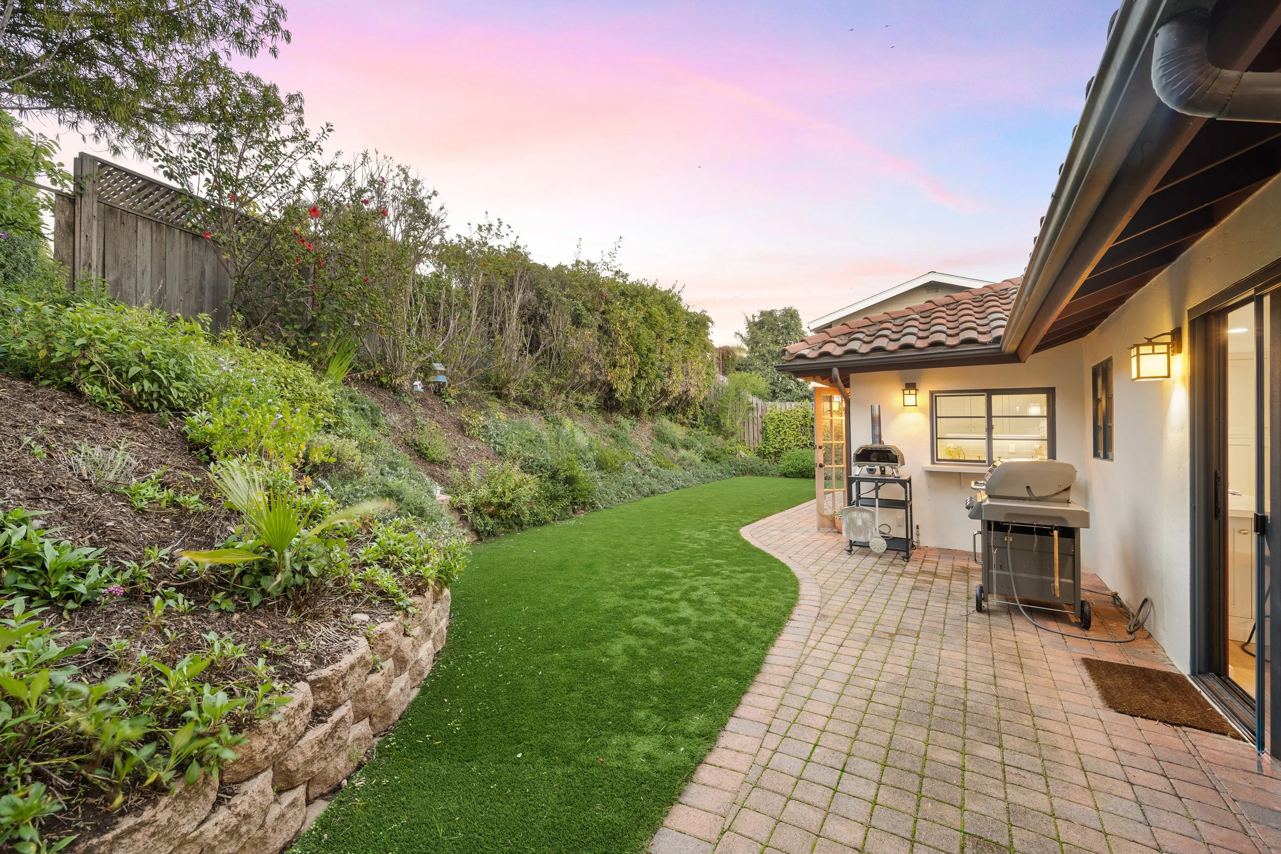 A backyard garden area with a brick patio, lush green grass, and garden bed on a slope with various plants and flowers. There is a white house with a tiled roof, outdoor lighting, and a barbecue grill, with a wooden fence and trees in the background, during dusk with a colorful sky.