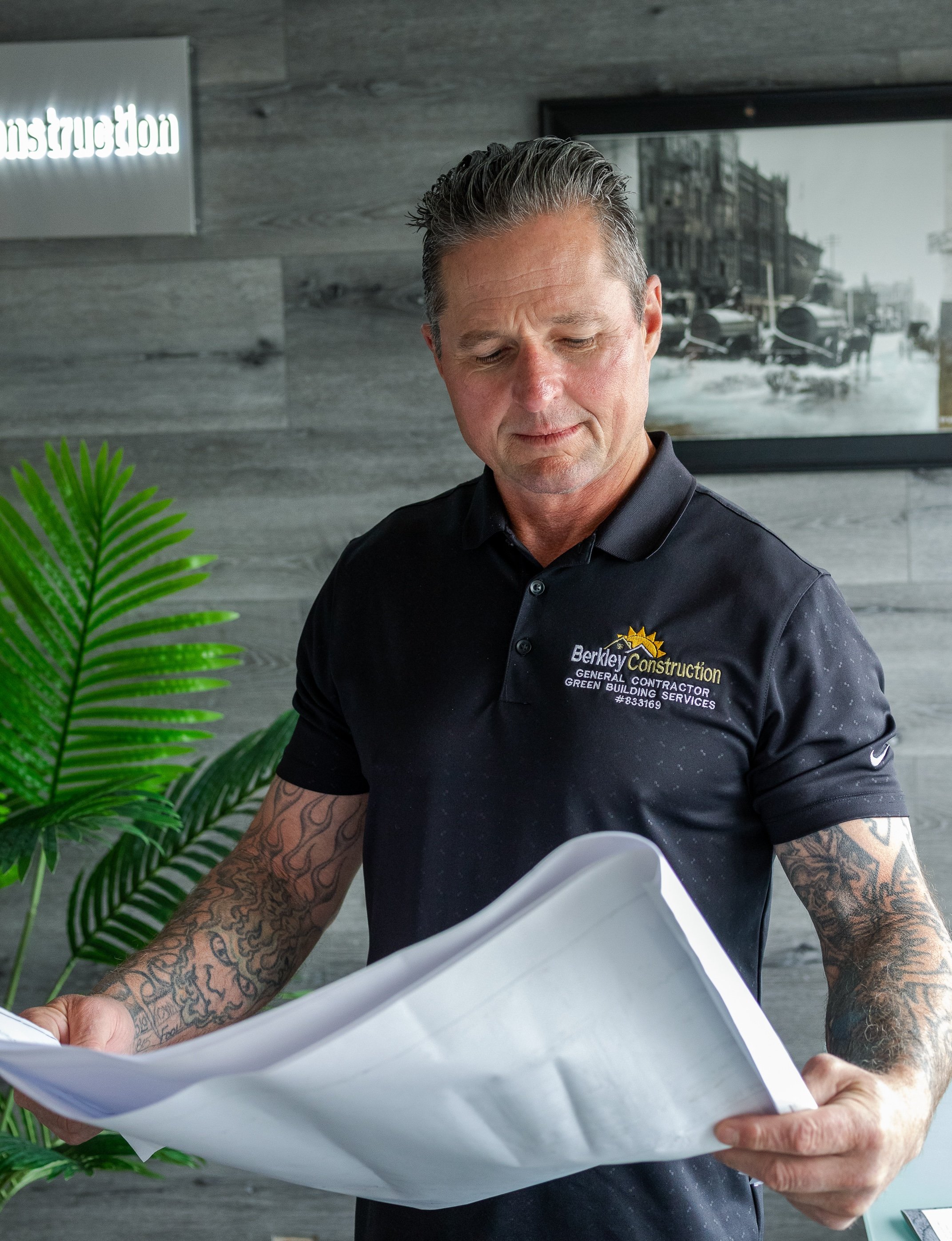 A man with gray hair and tattoos on his forearms, wearing a black polo shirt with a construction company's logo, looks down at a blueprint or construction plan. The background features a gray textured wall, a black and white cityscape photo, a green plant, and a sign that says 'Construction' in reverse.