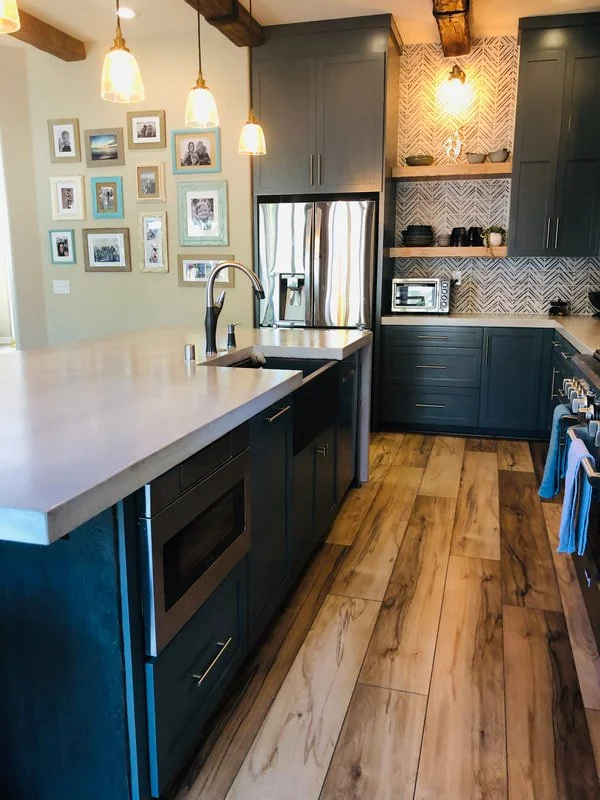 Modern kitchen with dark gray cabinets, stainless steel refrigerator, and light-colored countertop island with a sink. Open shelving with kitchenware and a built-in microwave, wood flooring, and pendant lighting.